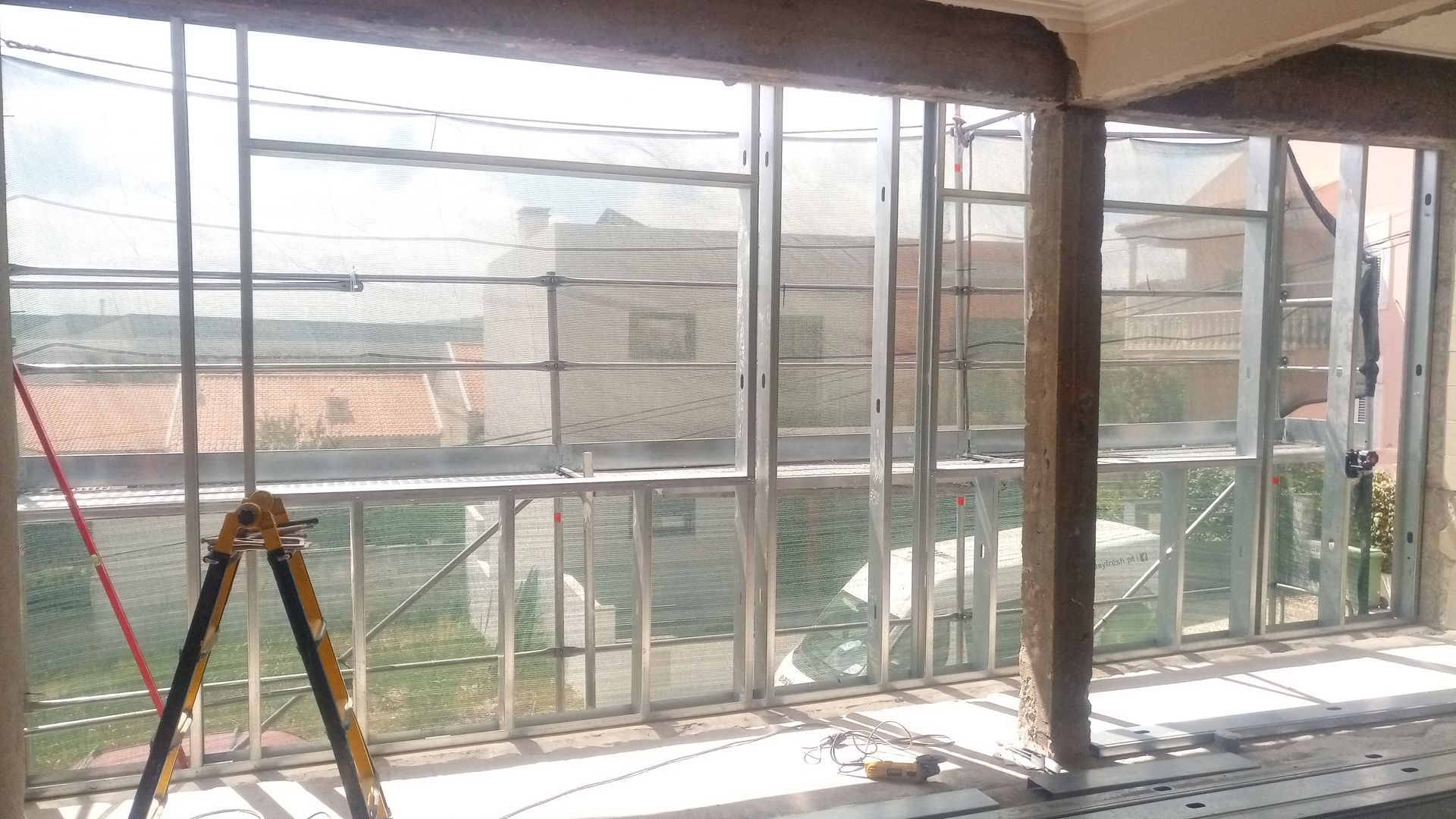 Vista interior de um edifício em construção, com janelas de vidro e estrutura metálica, com vista para uma área residencial.