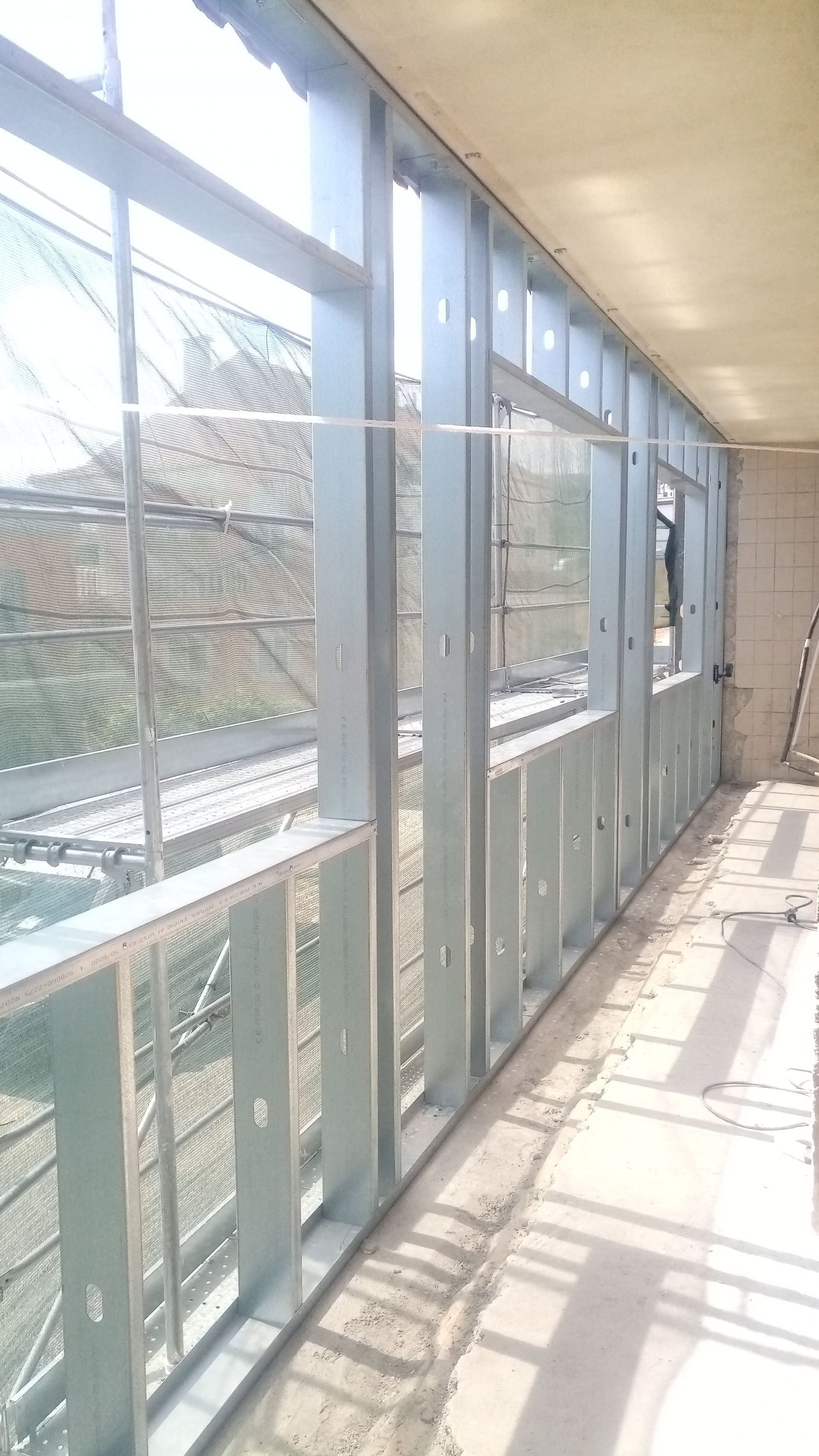 Estrutura metálica de uma parede em construção, com aberturas para janelas. Vista interna, com luz natural.