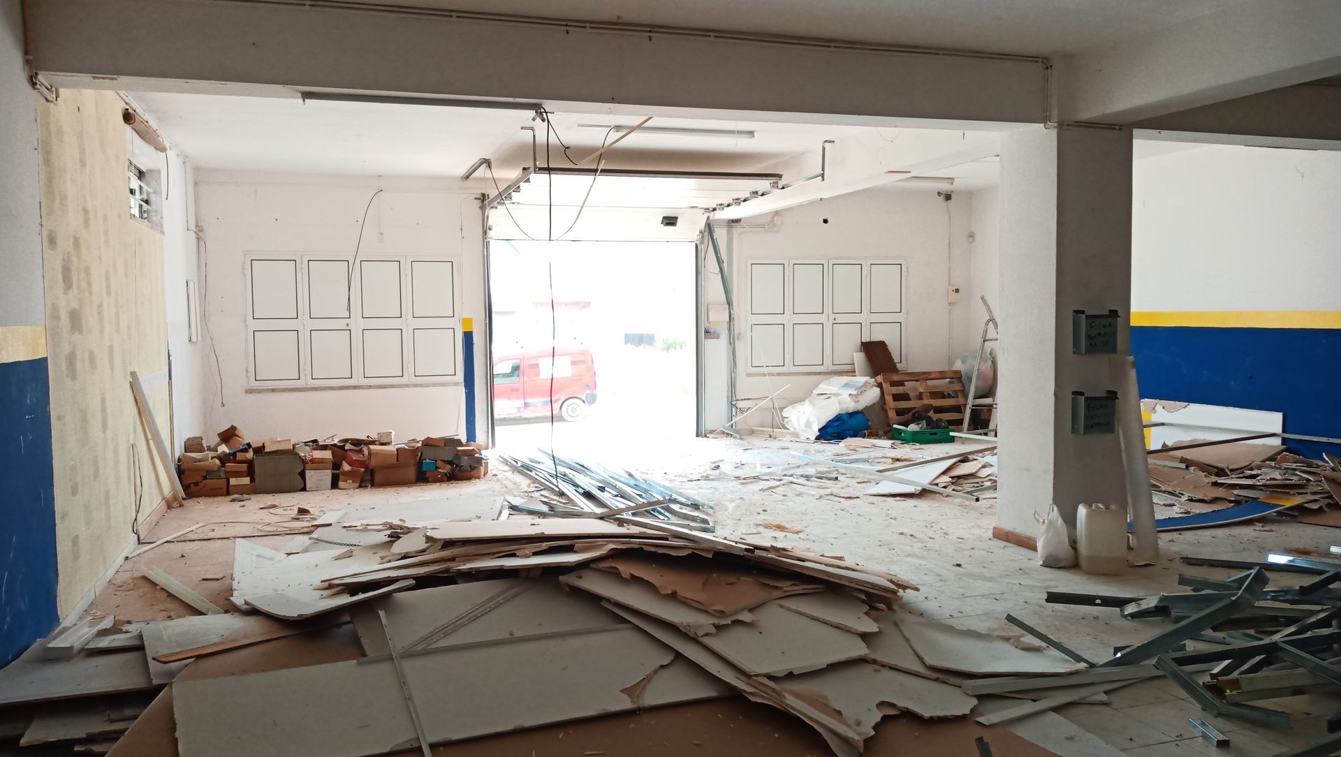 Interior de um edifício em ruínas, com detritos espalhados pelo chão. Uma abertura revela uma vista para o exterior.