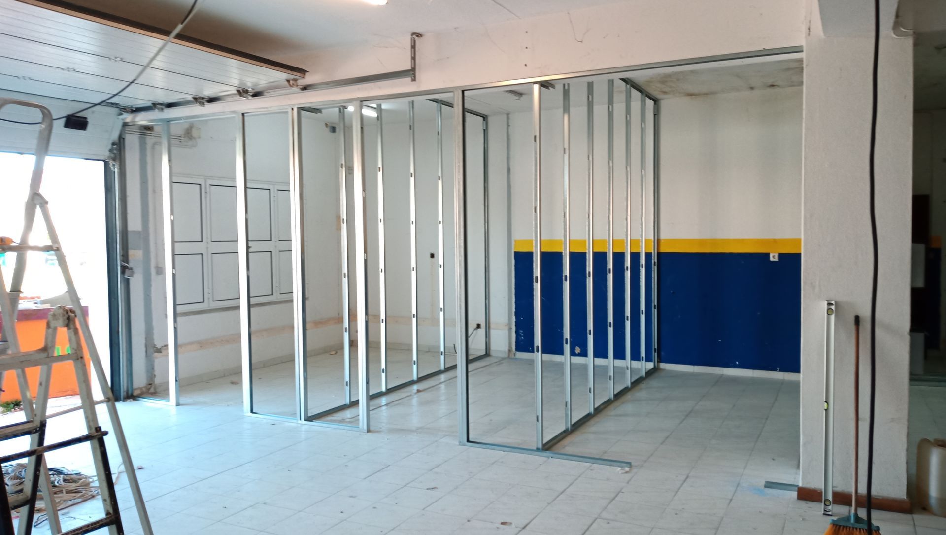 Estrutura metálica formando paredes internas em um cômodo em construção. As paredes estão parcialmente construídas, com piso de azulejo branco.