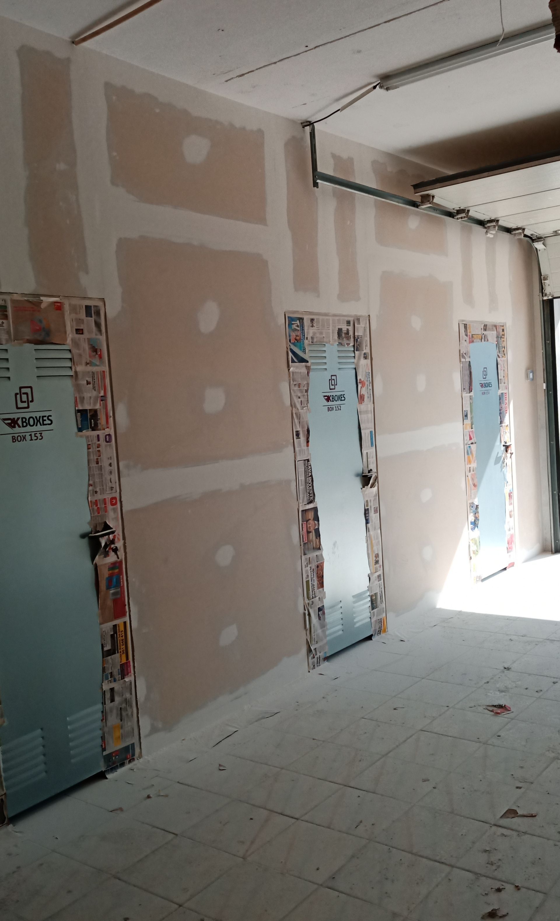 Corredor interno com parede de gesso inacabada e três portas de metal cobertas com jornal.