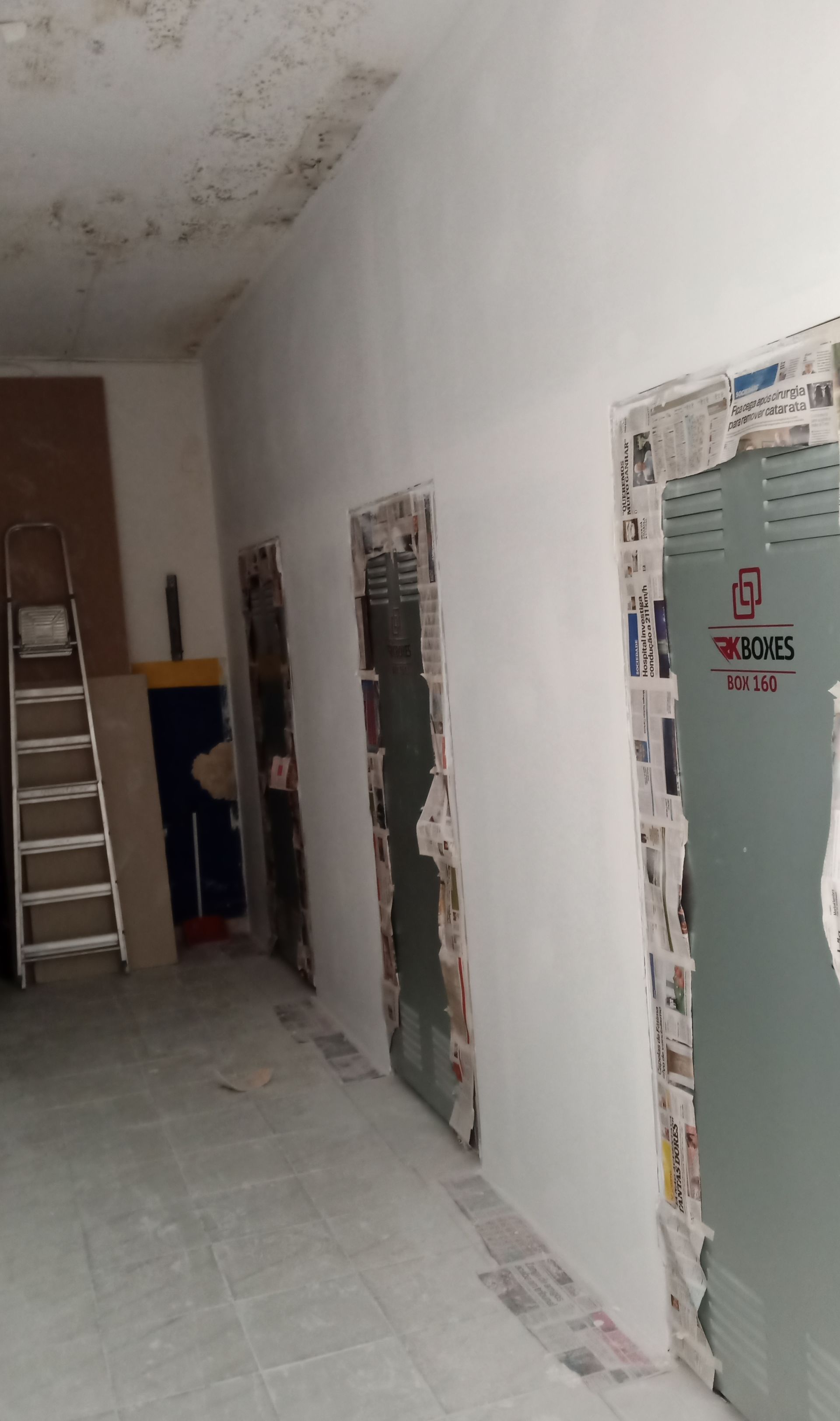 Corredor interno com três portas. As paredes são brancas e as portas estão cobertas com jornal. Uma escada está encostada na parede.