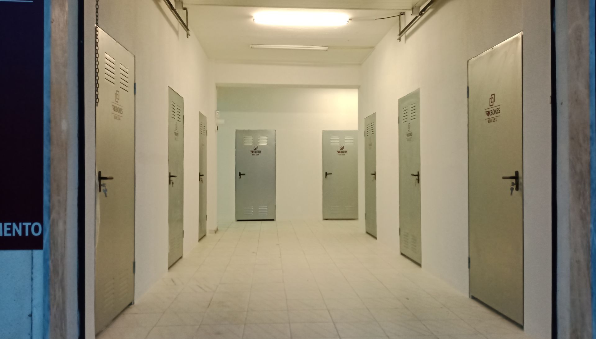 Um longo corredor com várias portas de metal cinza e paredes brancas sob luz fluorescente.