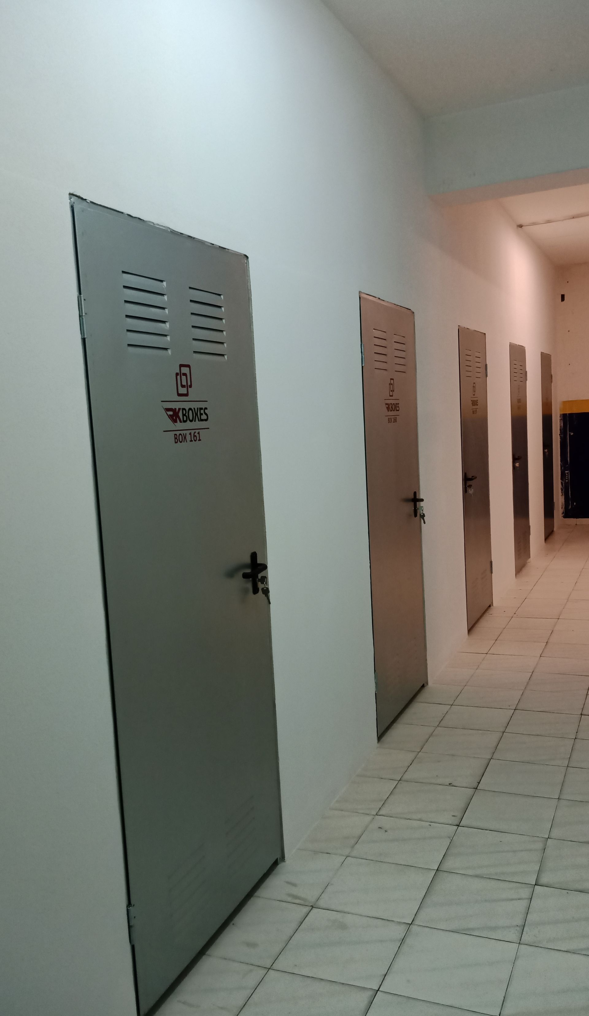Um corredor com várias portas de metal cinza para armários ao longo de uma parede branca, que leva a uma abertura escura.