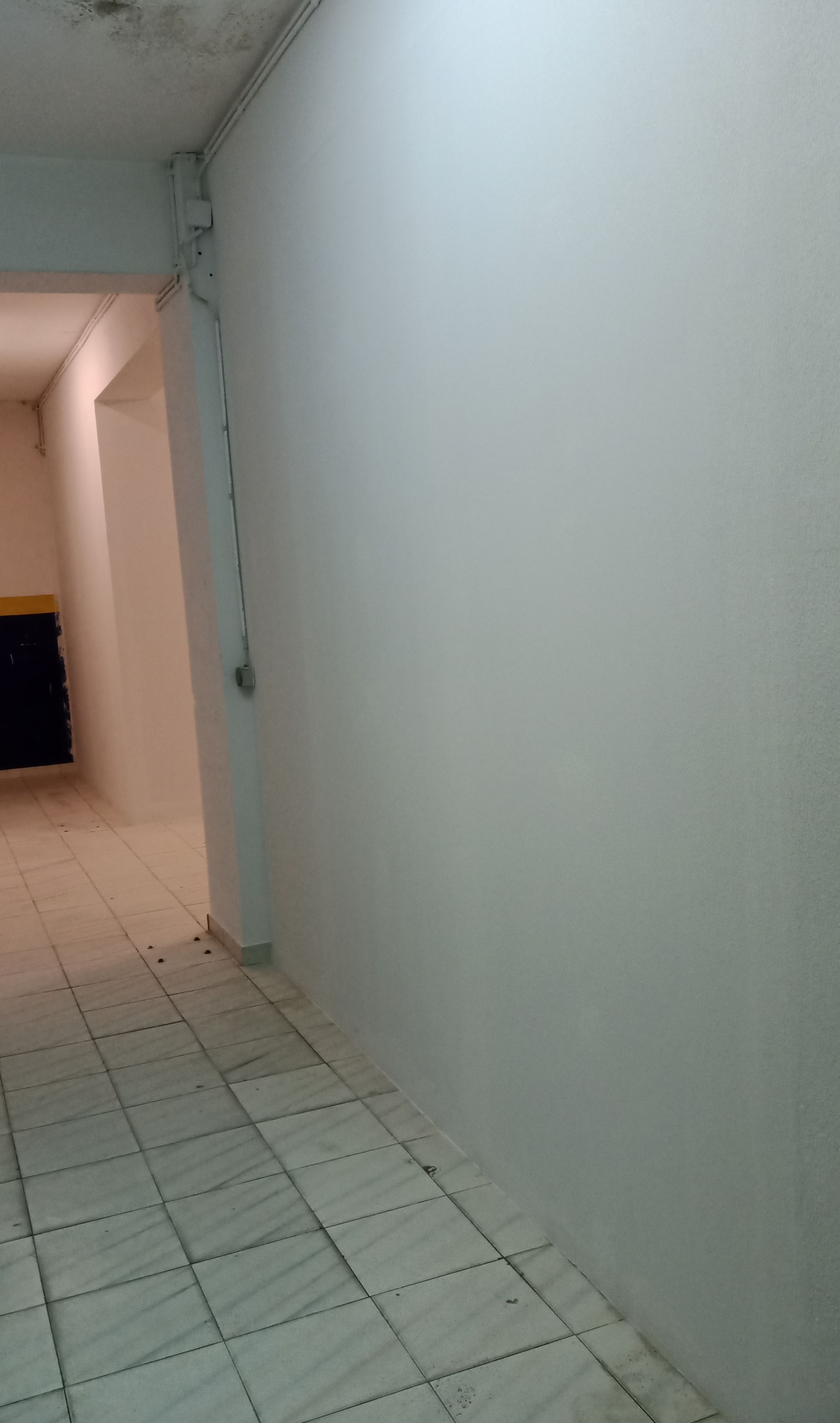 Corredor comprido com paredes brancas e piso de azulejo. Um espaço escuro é visível ao final.