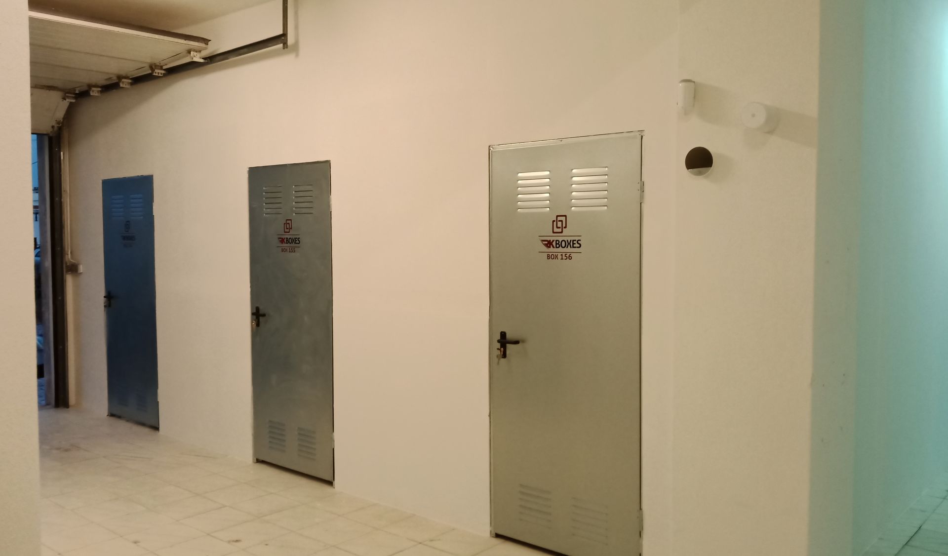 Um corredor com três portas de metal numeradas ao longo de uma parede branca; luzes fluorescentes no teto.
