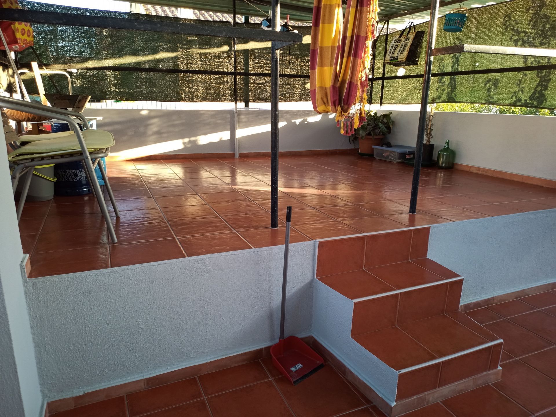 Terraço com piso de ladrilho vermelho, paredes brancas e escadas. Uma vassoura e uma rede estão presentes.