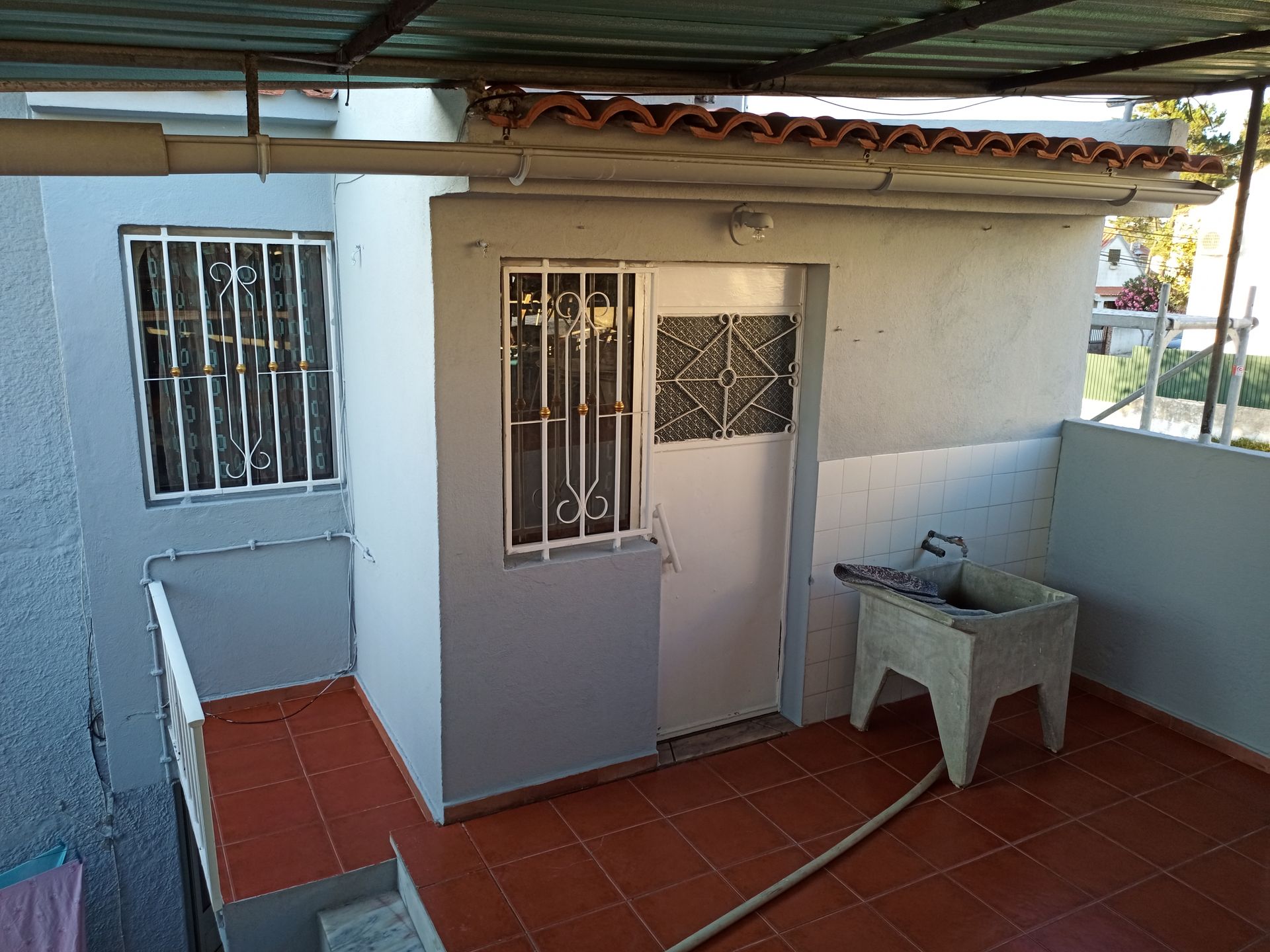 Pequeno cômodo externo com porta, janela gradeada e tanque de lavar roupa; piso de ladrilho vermelho.