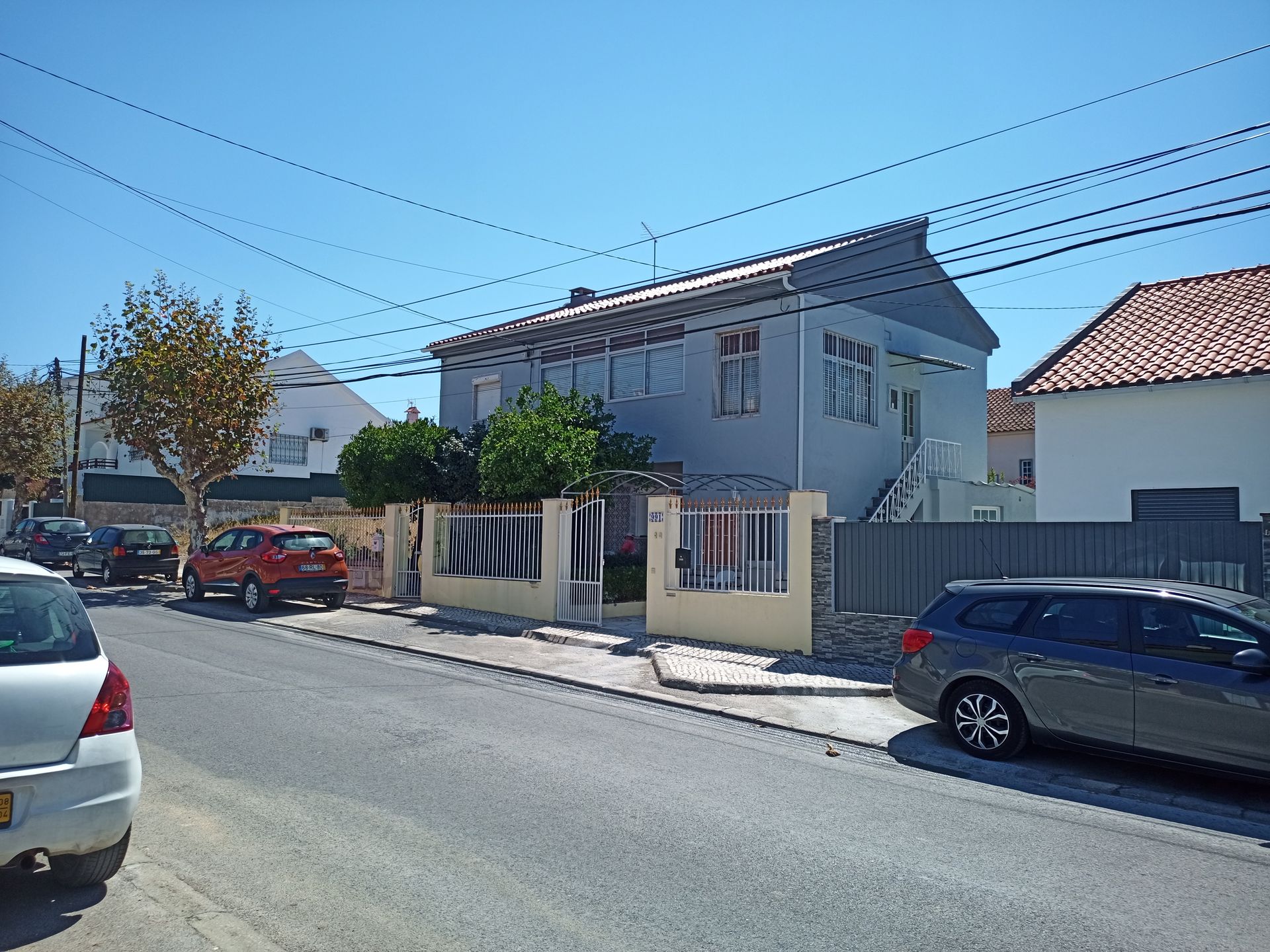 Prédio cinza de dois andares em uma rua com carros estacionados e fios de energia; céu azul.