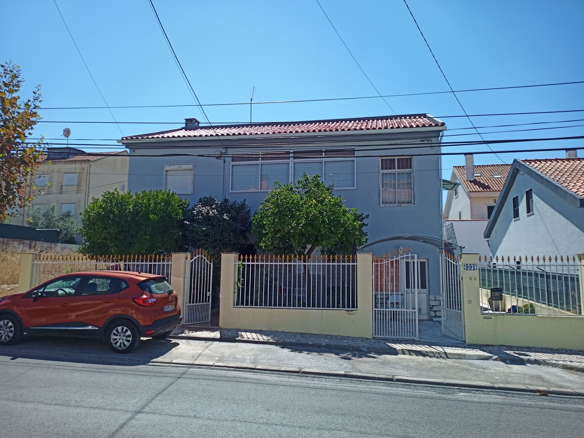 Casa azul de dois andares atrás de uma cerca amarela, com um carro laranja estacionado em frente em um dia ensolarado.