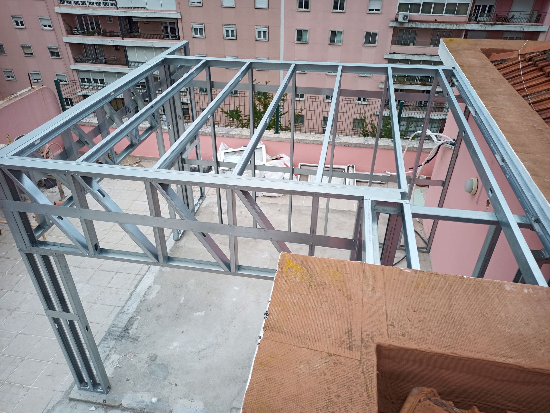 Cobertura com estrutura metálica em construção em um telhado com prédio rosa ao fundo.