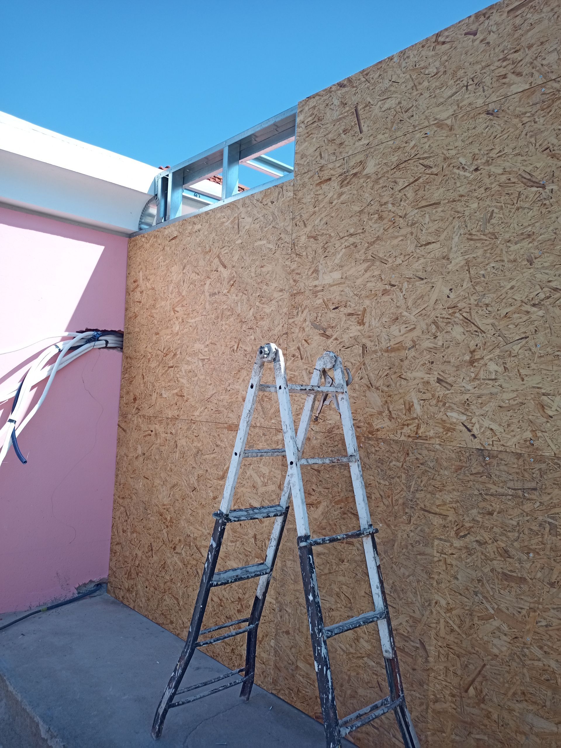 Escada encostada em uma parede inacabada de OSB, com estrutura metálica acima. Parede rosa à esquerda.