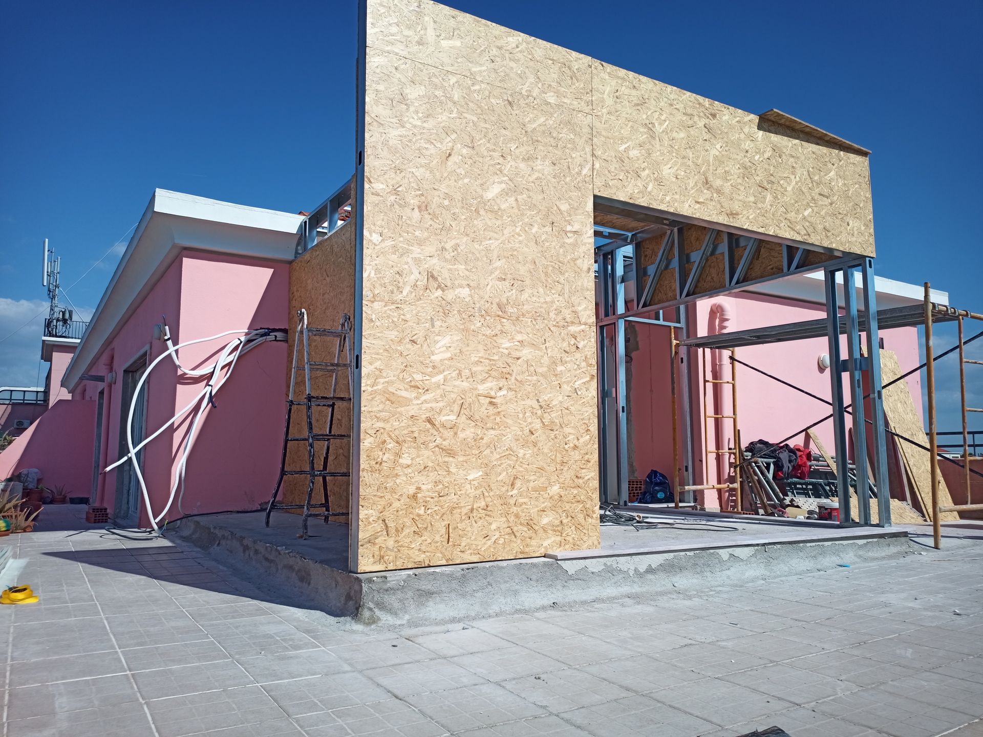 Obras no telhado de um prédio rosa; painéis de madeira compensada e estrutura metálica são visíveis. Céu azul ao fundo.