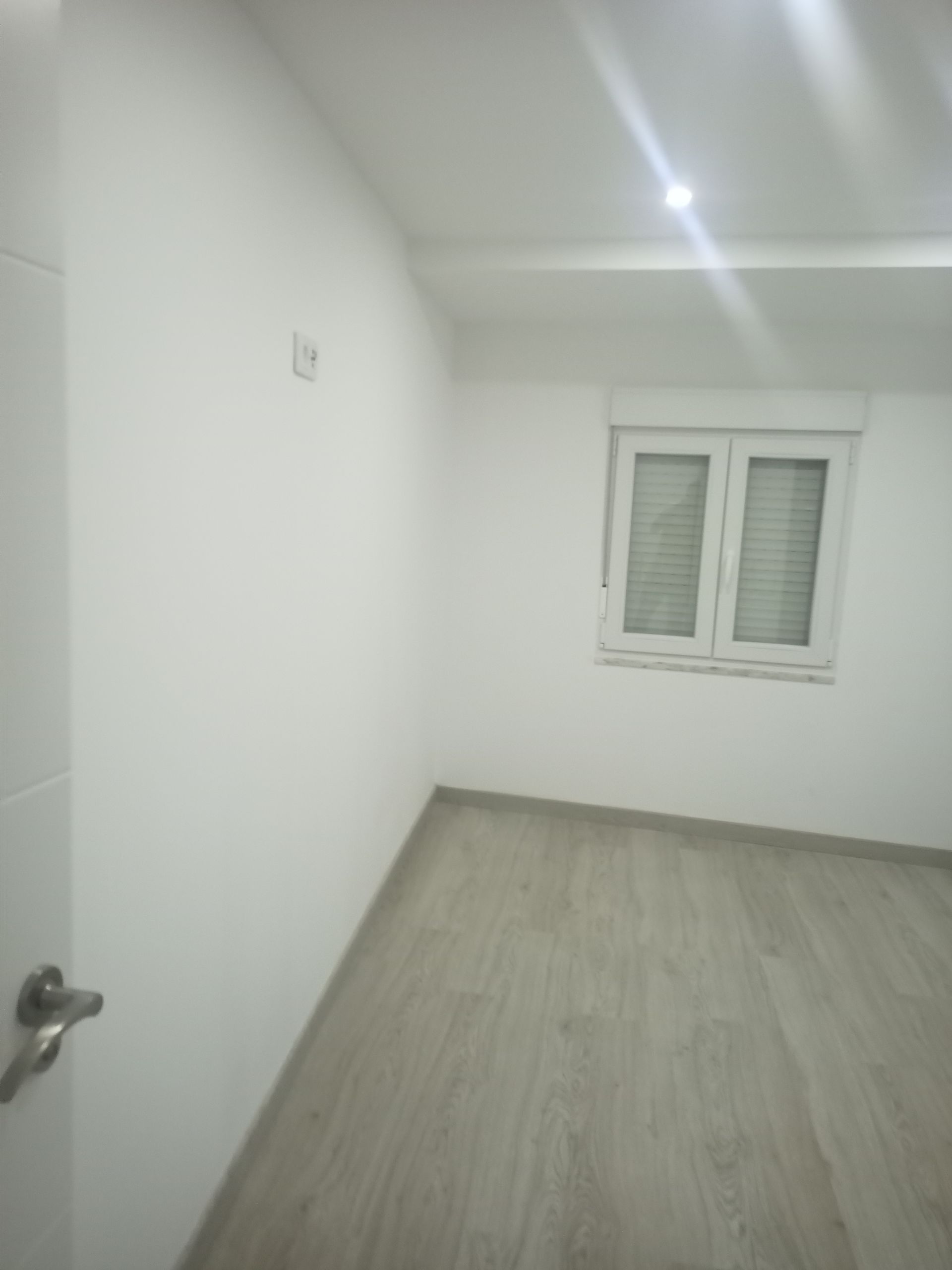 Quarto vazio, com paredes brancas, janela, piso que imita madeira clara e uma porta à esquerda.