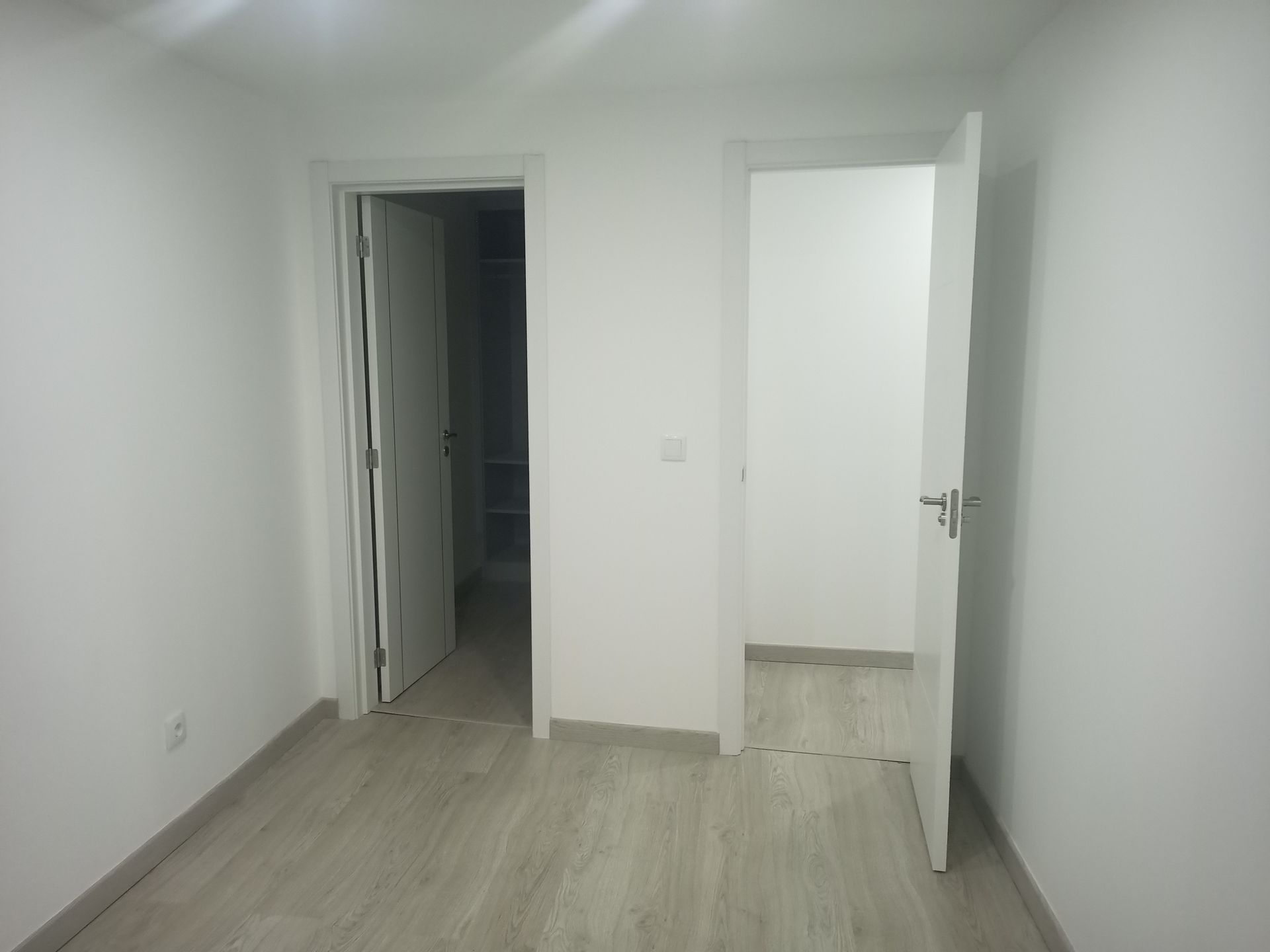 Quarto vazio com duas portas brancas abertas, piso que imita madeira clara e paredes brancas.