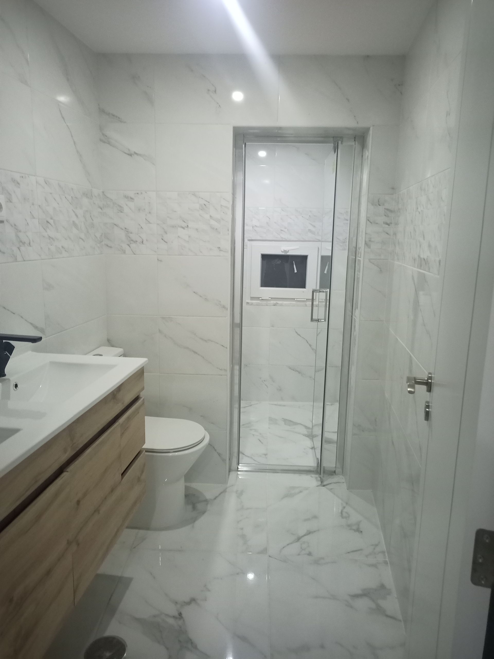 Banheiro moderno com azulejos de mármore branco, bancada de madeira e box de vidro.