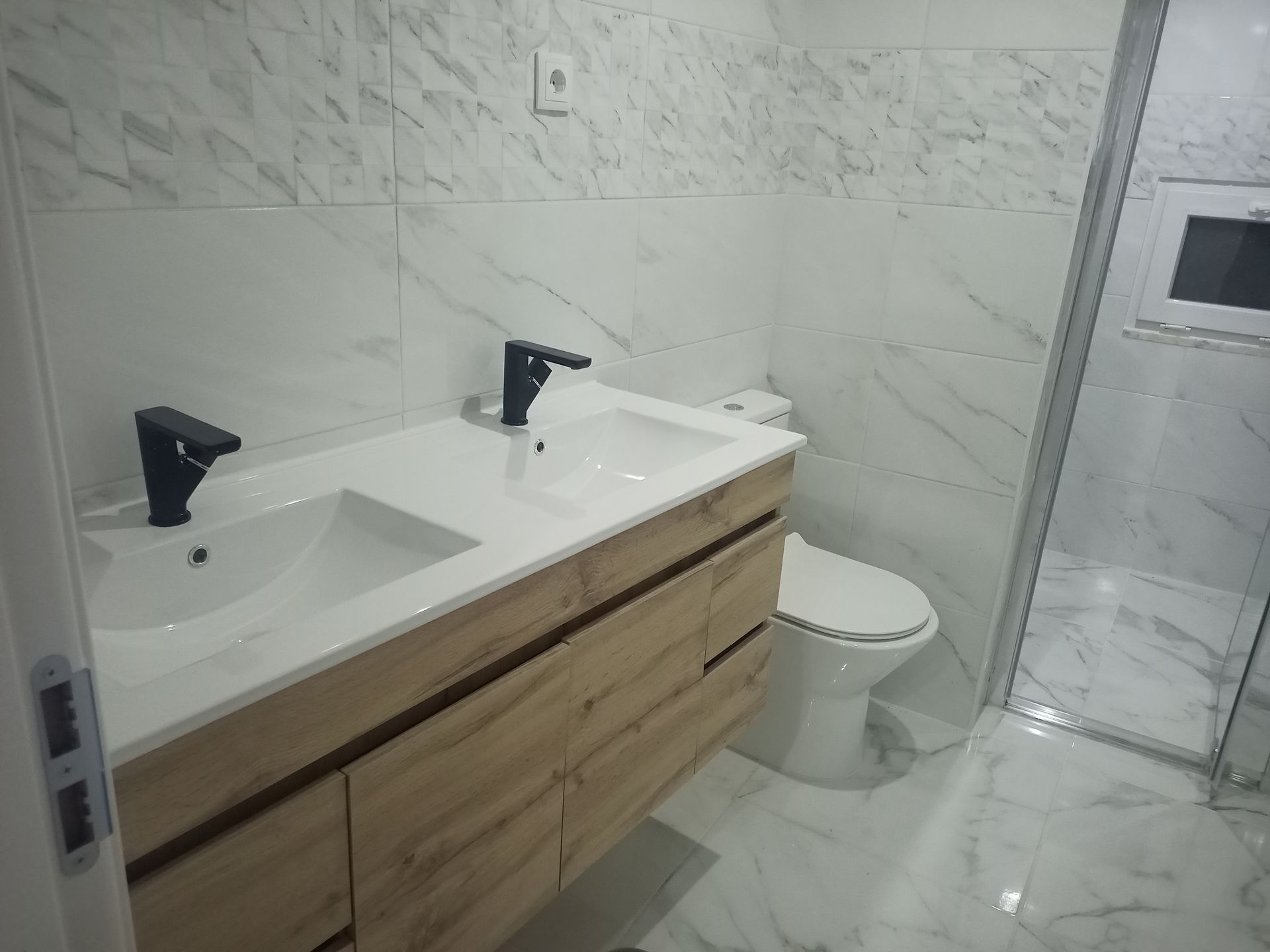 Banheiro com azulejos imitando mármore, pia dupla com torneiras pretas e vaso sanitário. O chuveiro é visível à direita.