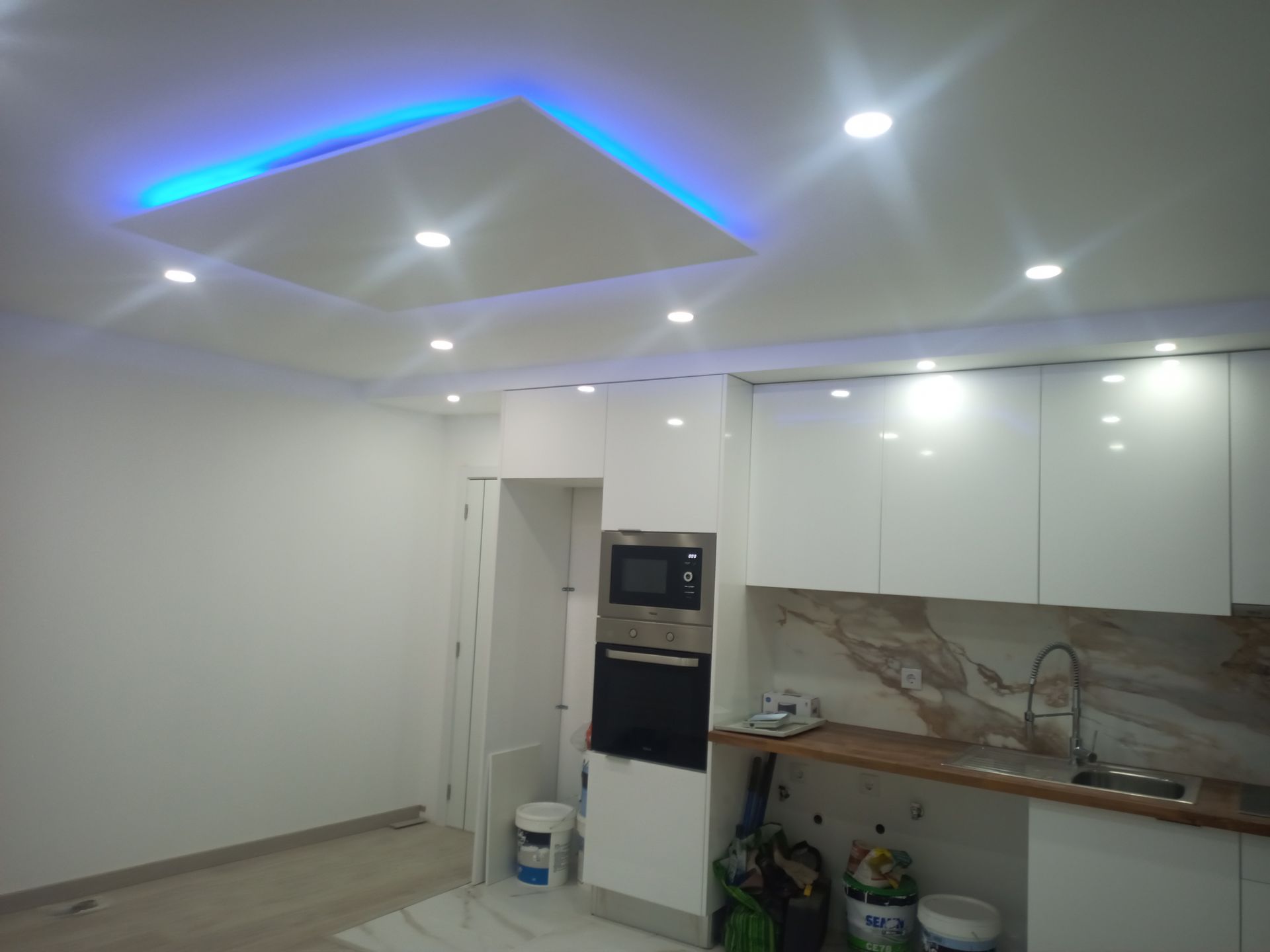 Cozinha moderna branca com iluminação embutida, detalhe azul no teto e eletrodomésticos embutidos.