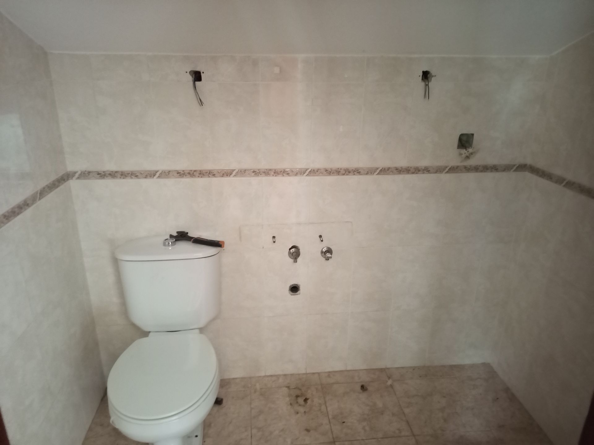 Banheiro vazio com vaso sanitário. Paredes revestidas com azulejos bege, faixa marrom, encanamento aparente.