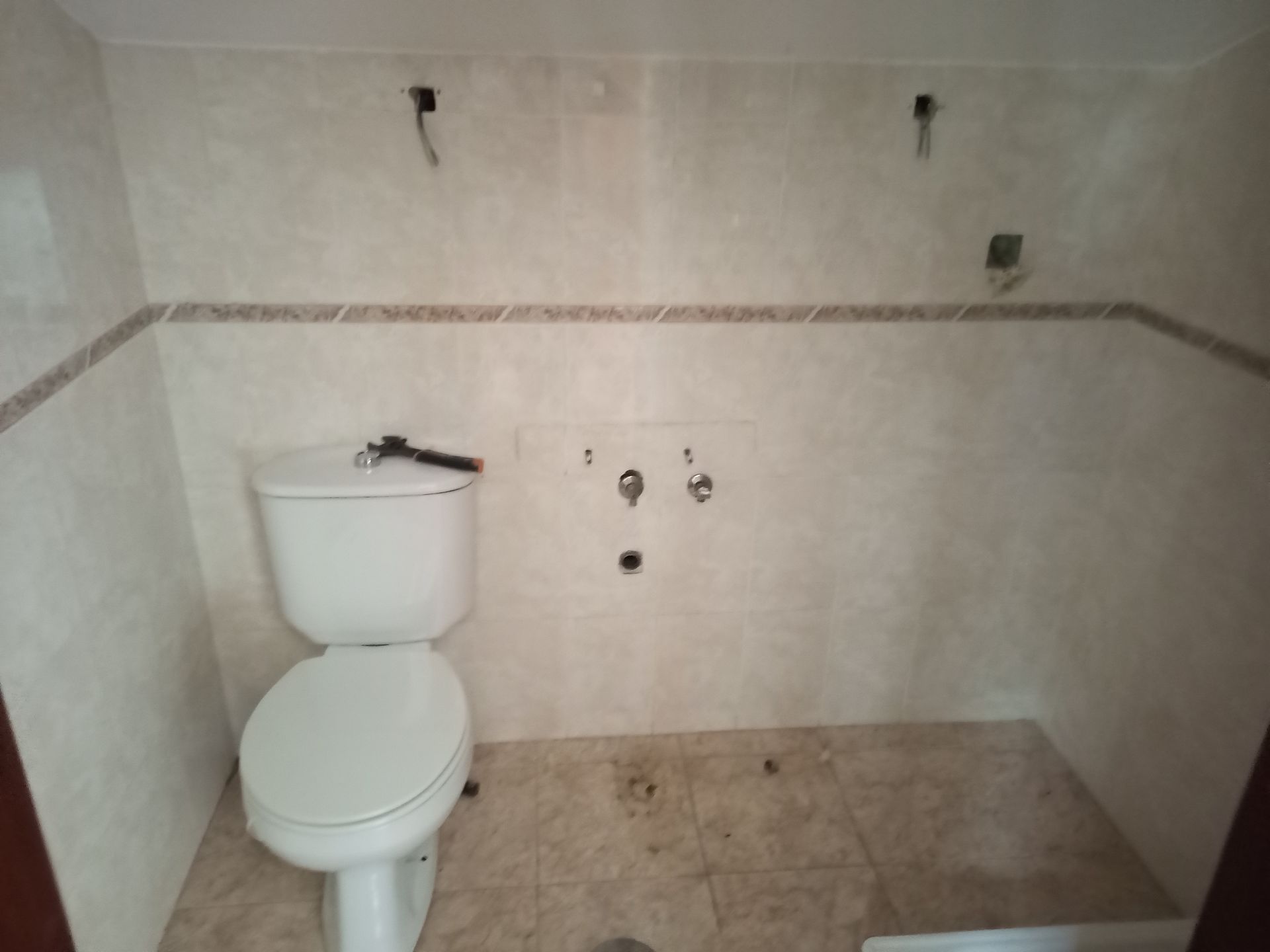Banheiro com vaso sanitário instalado. Encanamento aparente na parede. Azulejos e acabamentos bege.