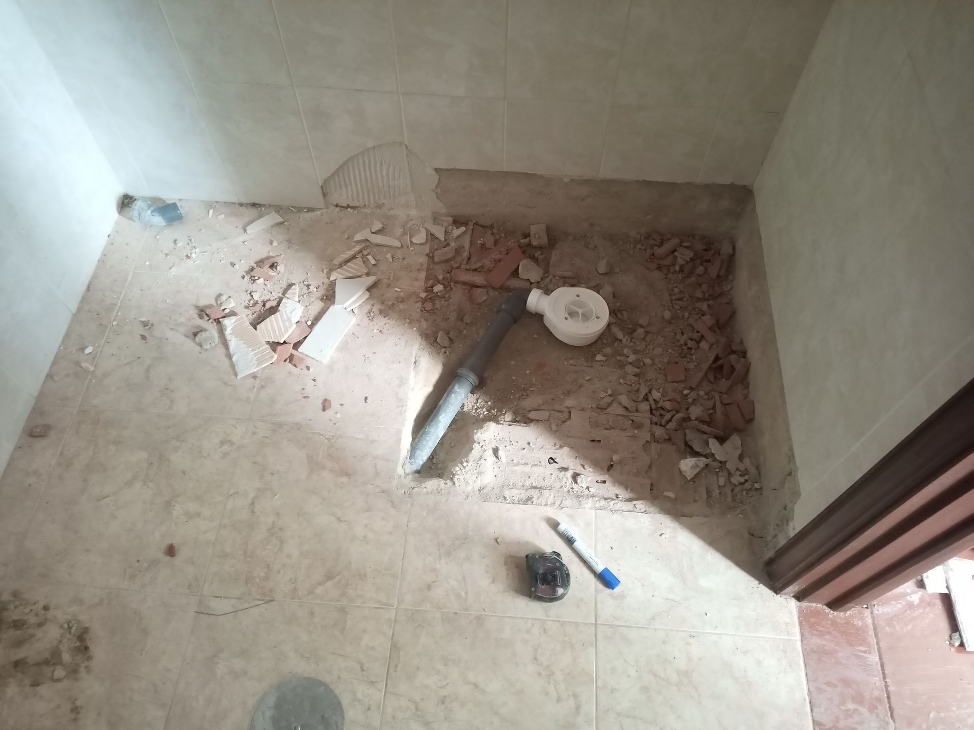 Piso de banheiro demolido, mostrando encanamento e entulho. As paredes são brancas e bege; uma porta é visível.