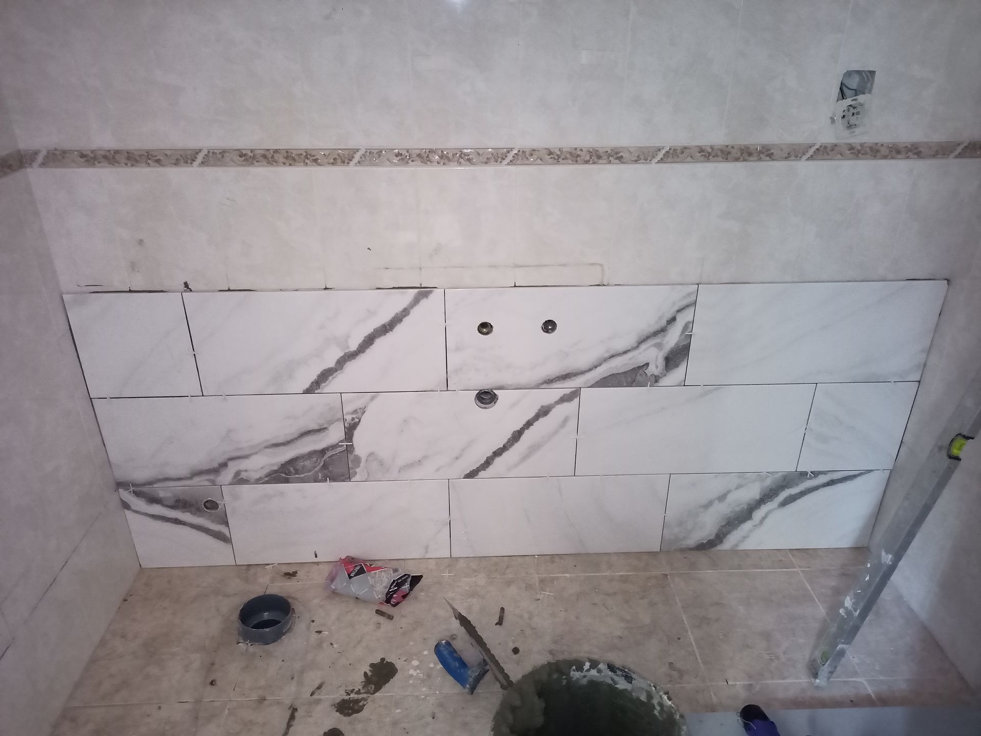 Parede do banheiro em construção, parcialmente revestida com azulejos imitando mármore; instalações hidráulicas visíveis; ferramentas e entulho no chão.