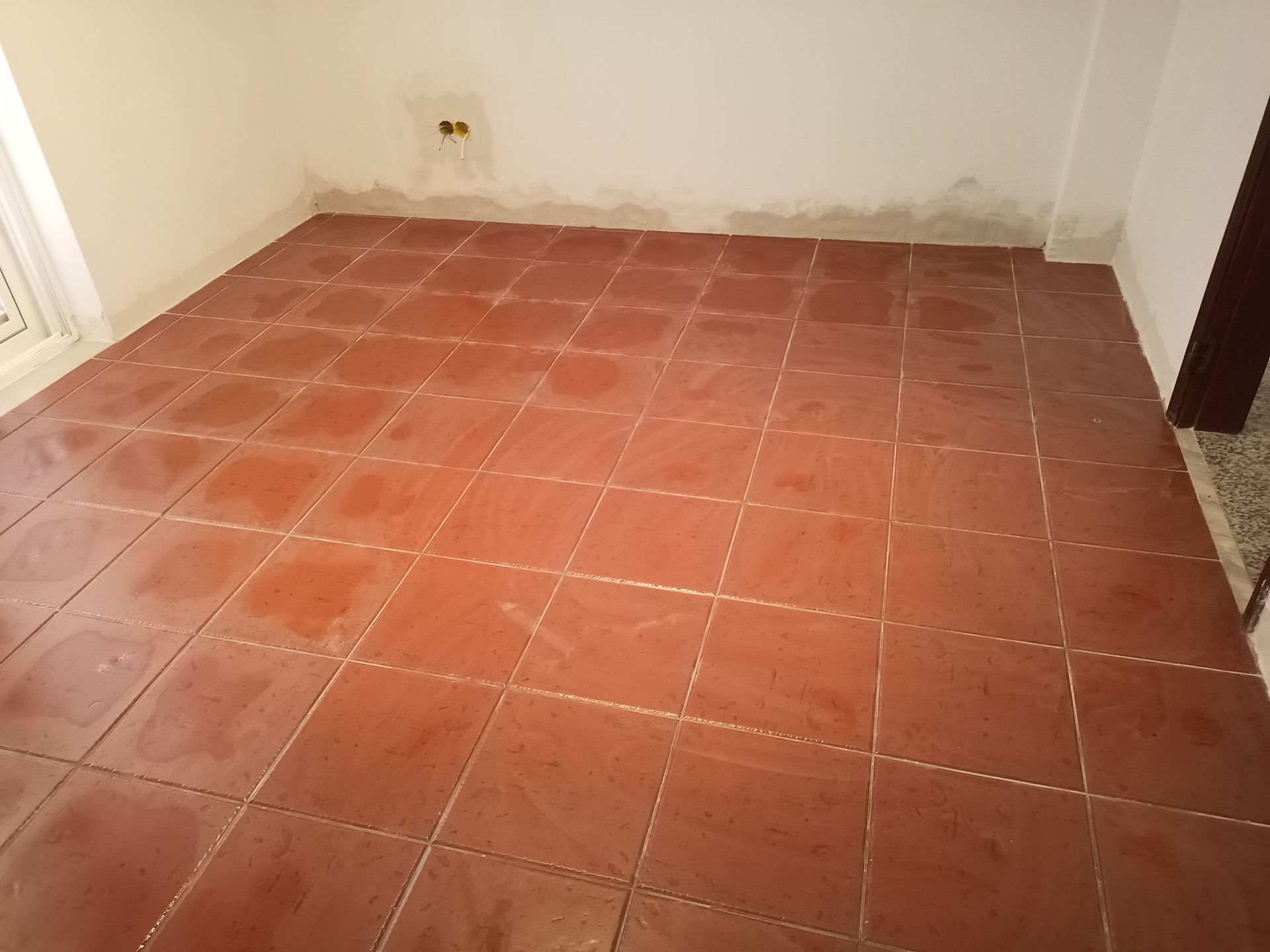Piso de ladrilho vermelho em um quarto com paredes brancas, uma porta e um interruptor de luz.