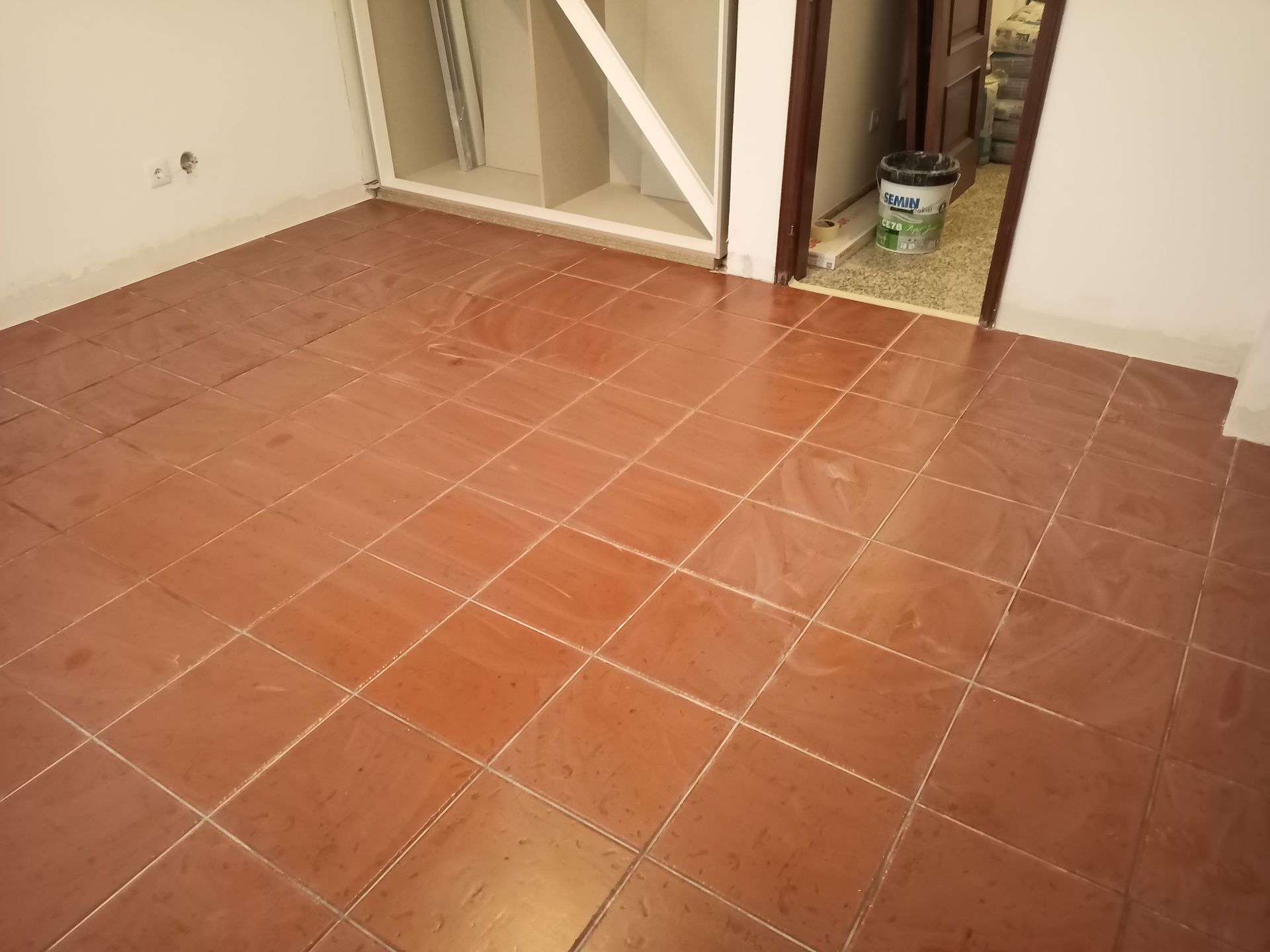 Piso de azulejos marrons em um cômodo, com uma porta parcialmente visível e prateleiras embutidas.