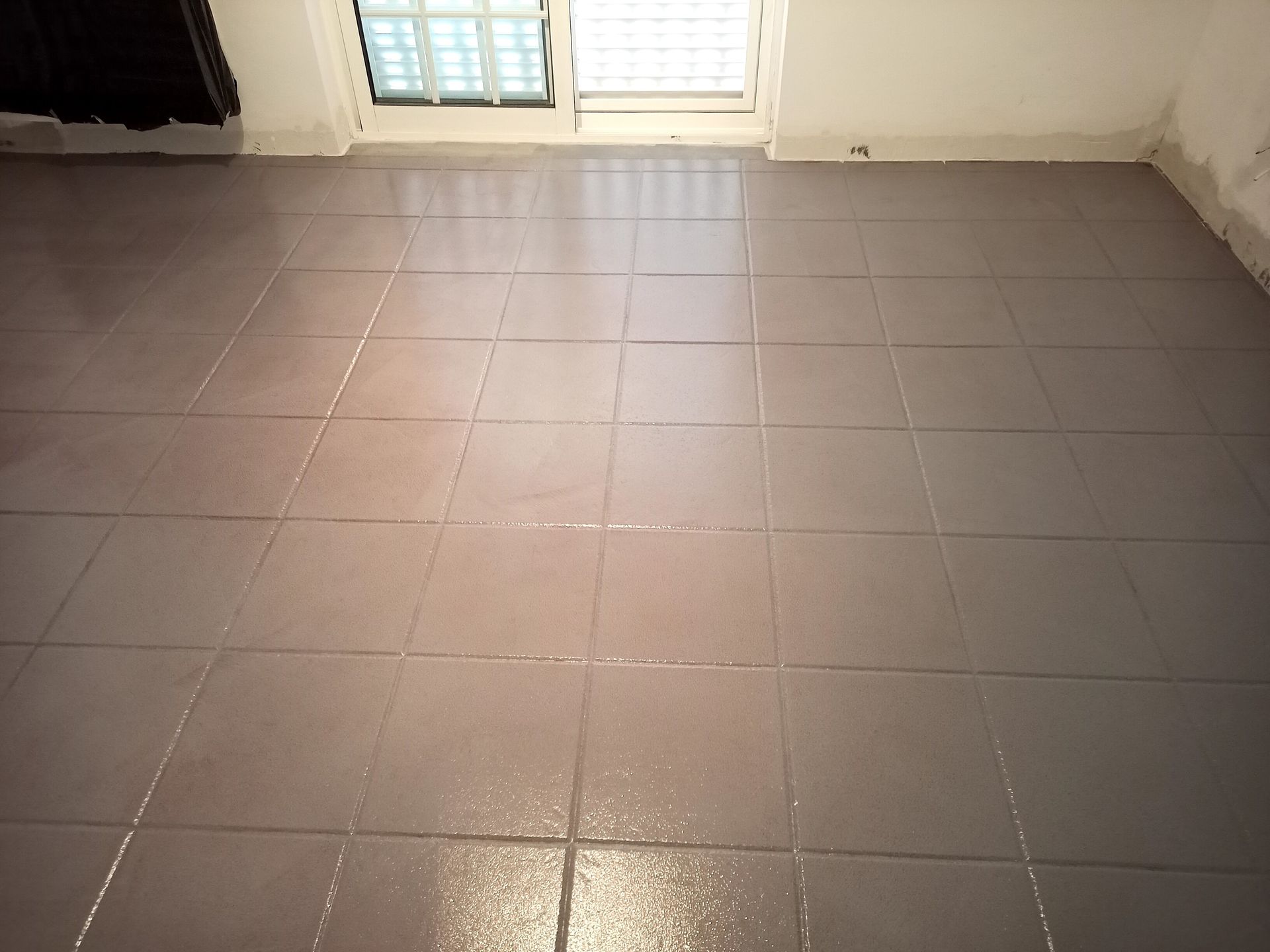 Piso de azulejos cinza em um cômodo, com uma janela e paredes brancas visíveis ao fundo.