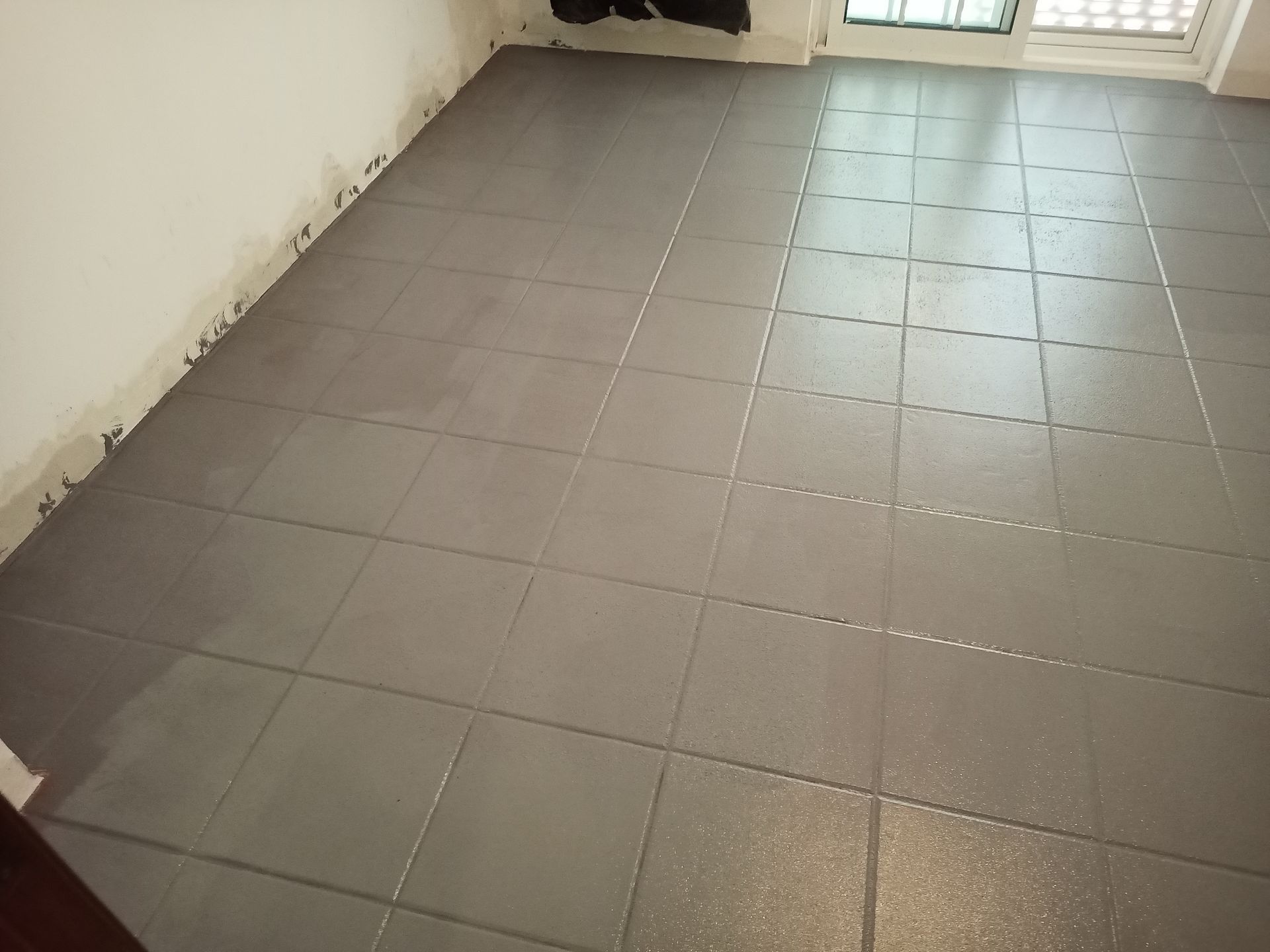 Piso de azulejos cinza, rejuntado recentemente, em um cômodo com paredes brancas e uma janela.