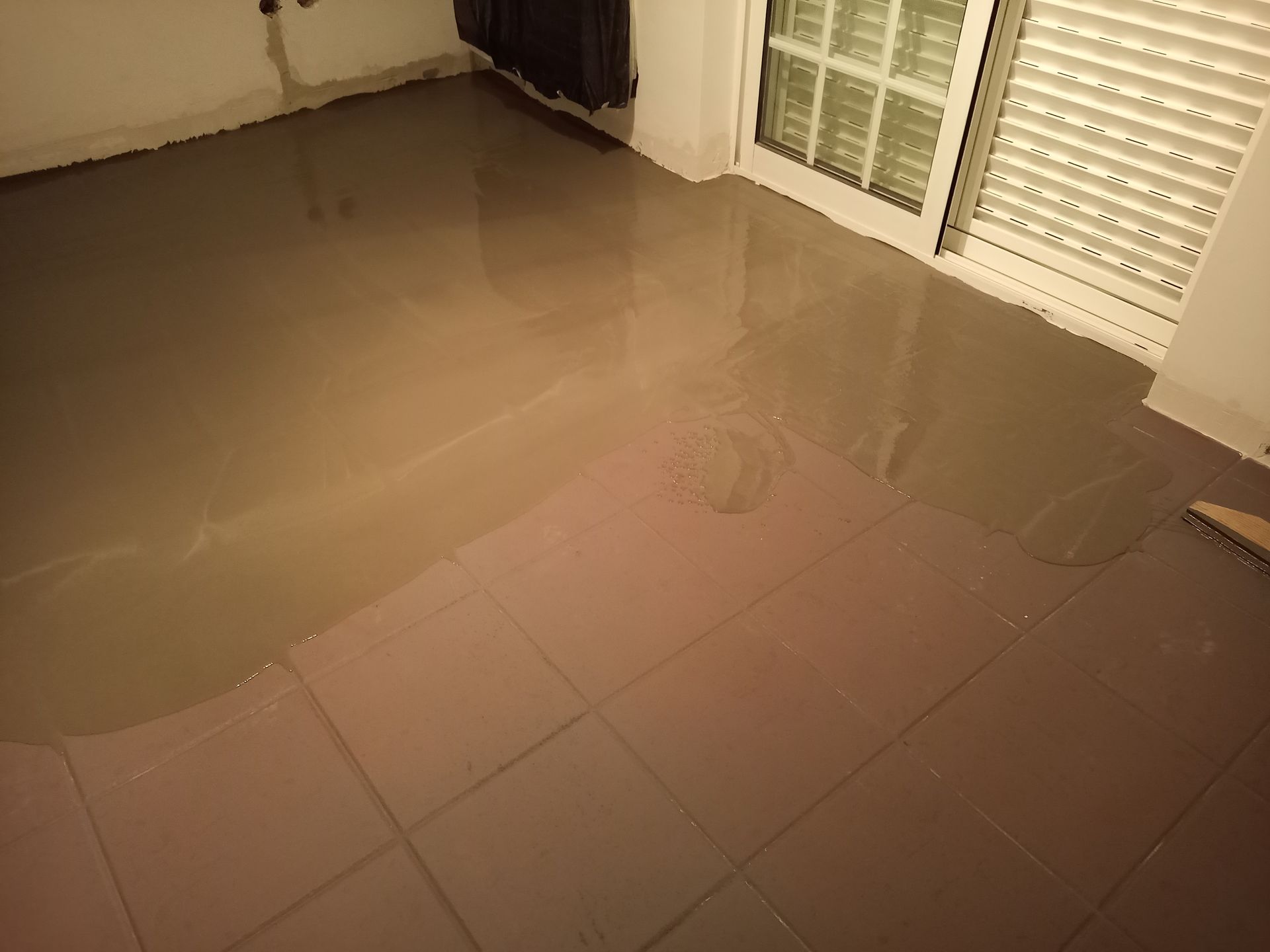 Piso autonivelante recém-instalado em um cômodo. Revestimento líquido marrom em placas de espuma rosa, próximo a uma porta de correr fechada.