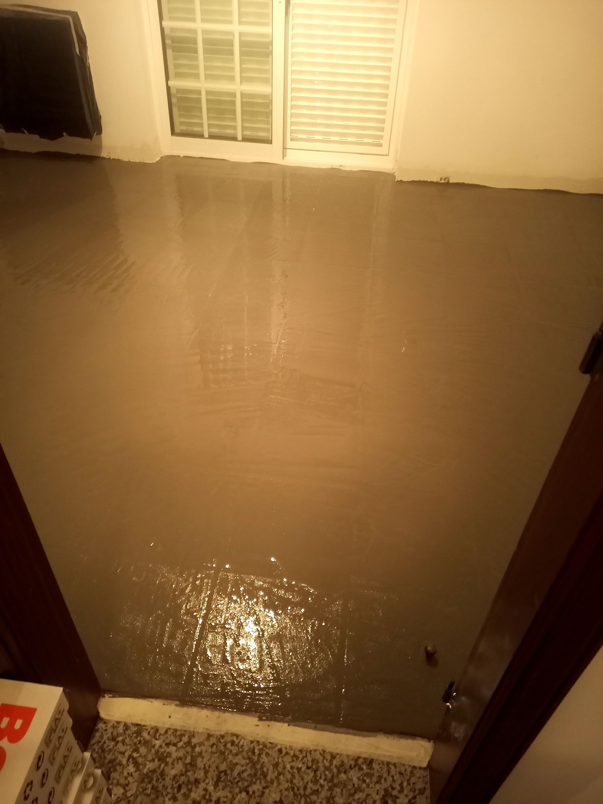Piso de concreto cinza, fresco e úmido, refletindo a luz em um cômodo. Porta/janela branca ao fundo.