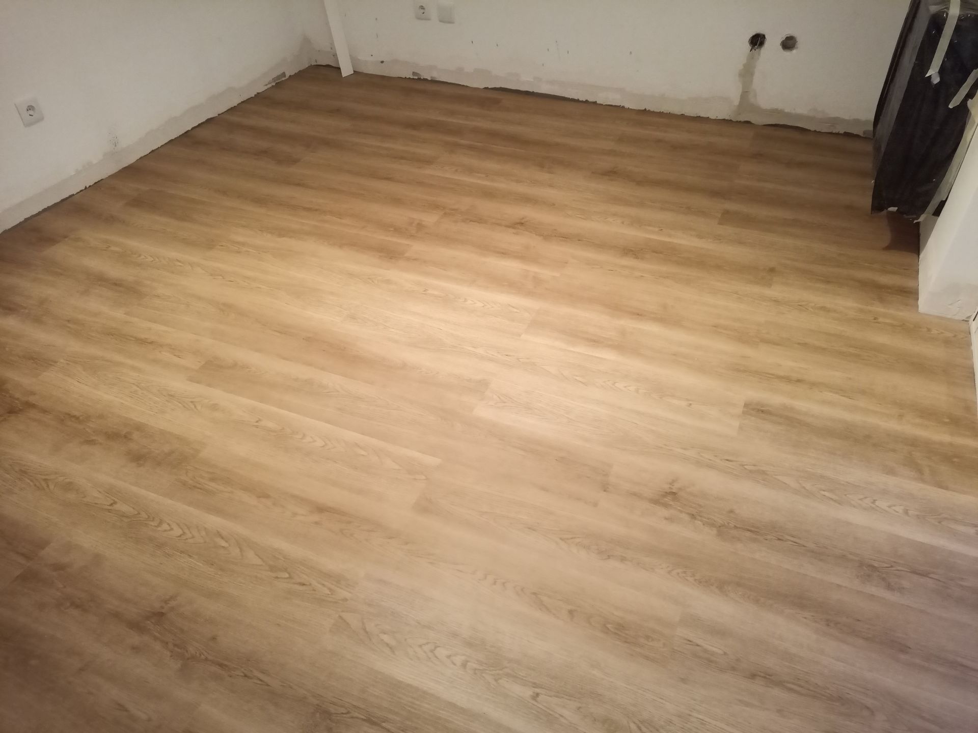Quarto com piso de madeira, paredes brancas e tomadas elétricas visíveis.