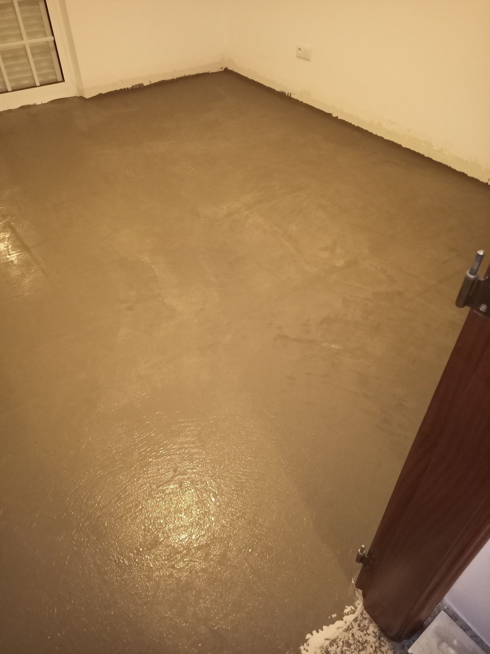 Sala com piso de concreto recém-moldado, refletindo a luz ambiente. Piso marrom, paredes brancas.