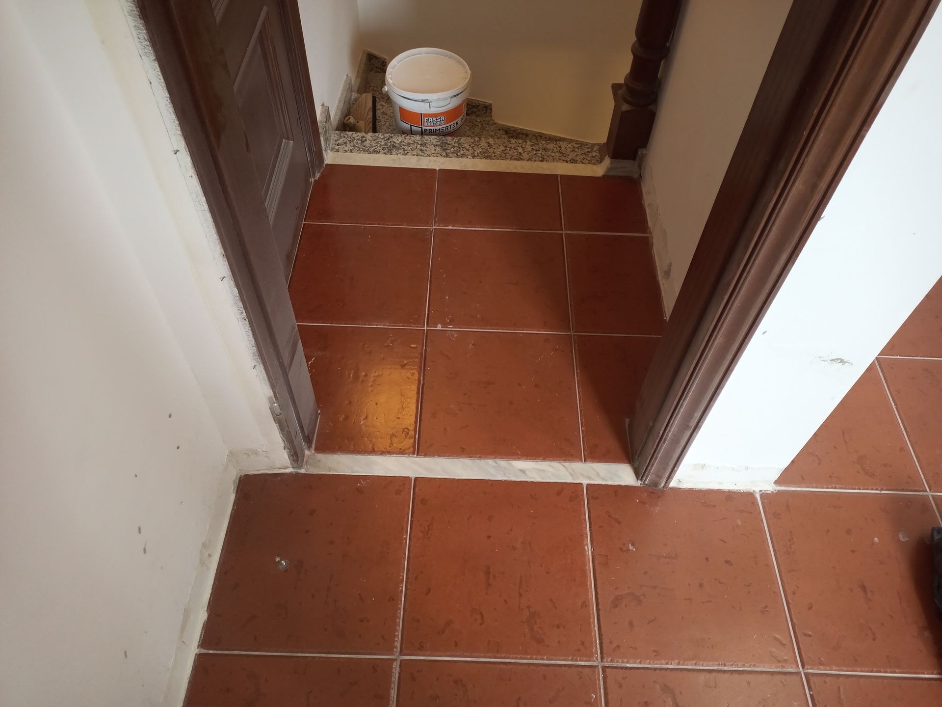 Piso de ladrilho marrom na entrada de uma porta. Um balde parcialmente visível está ao fundo.