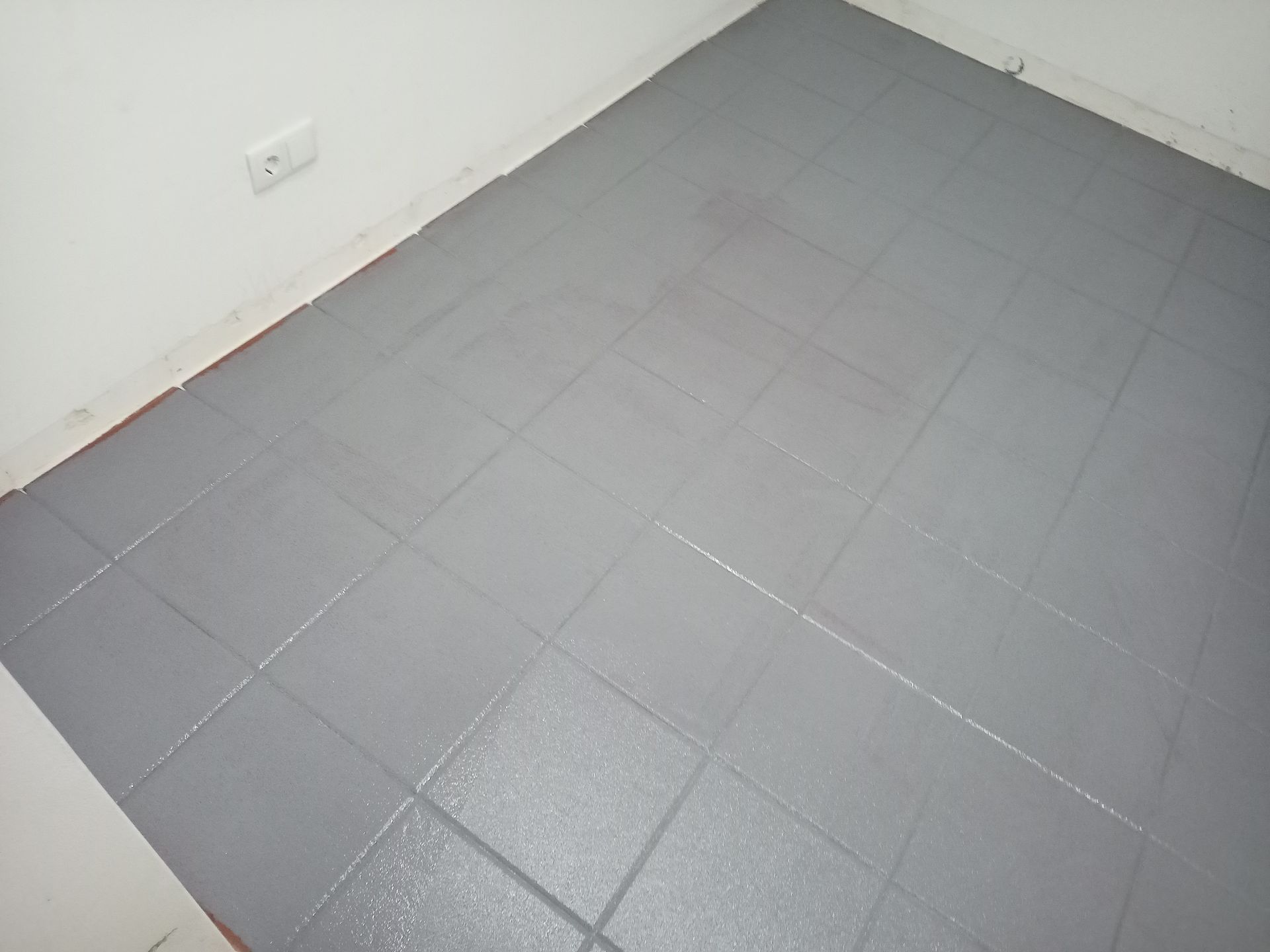 Piso de azulejos pintado de cinza em um cômodo, com uma parede branca e tomada elétrica visível.