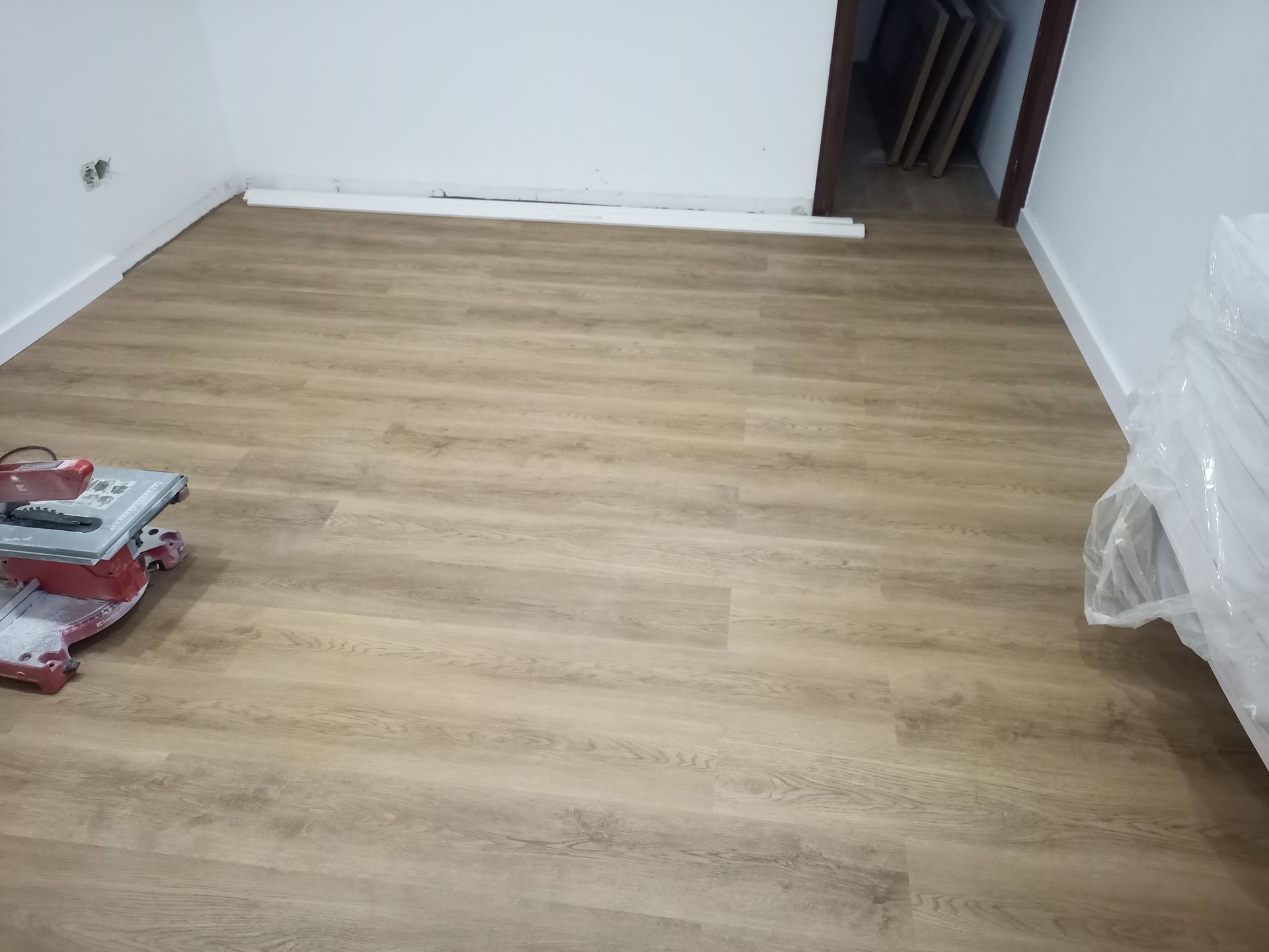 Sala com piso imitando madeira em tom castanho claro sendo instalado. Uma serra e materiais embalados estão presentes.