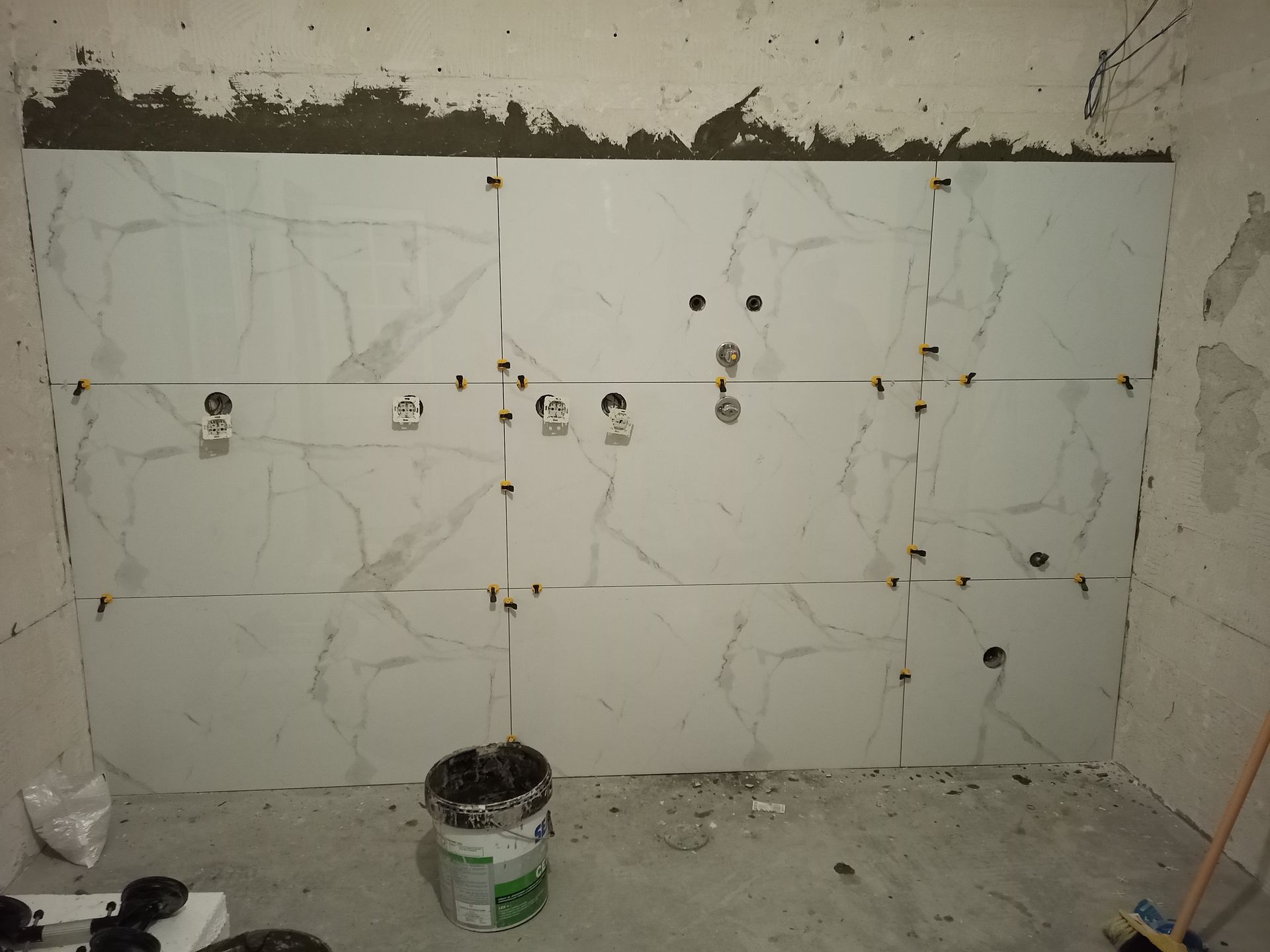 Parede com grandes ladrilhos brancos imitando mármore, alguns parcialmente instalados com espaçadores, cena de construção.