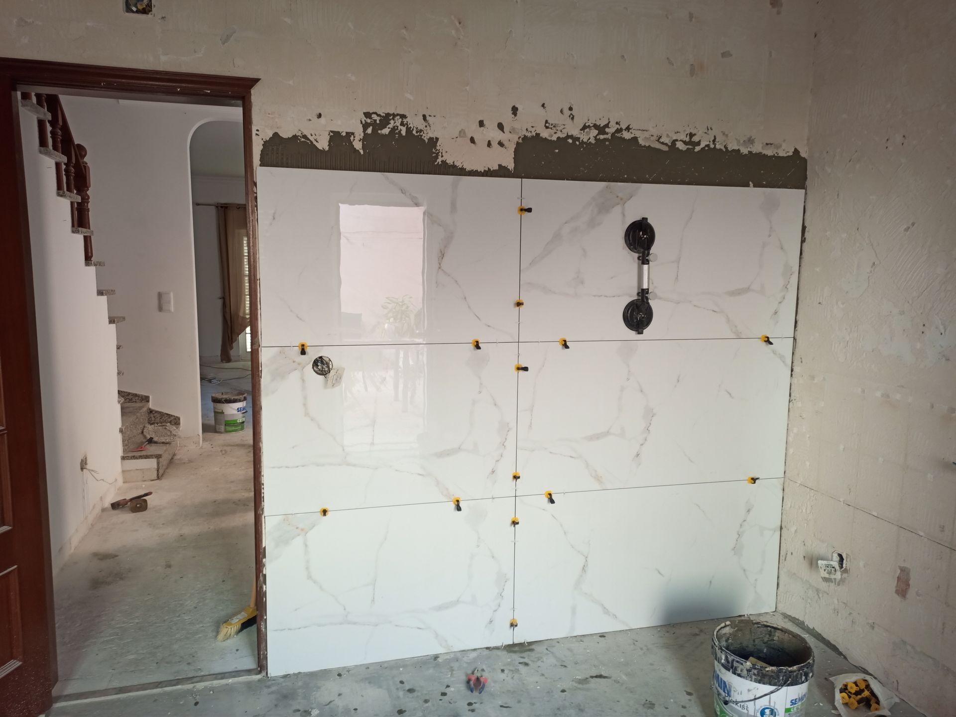 Parede parcialmente revestida com azulejos imitando mármore, ventosa fixada, porta à esquerda, balde e escada visíveis.