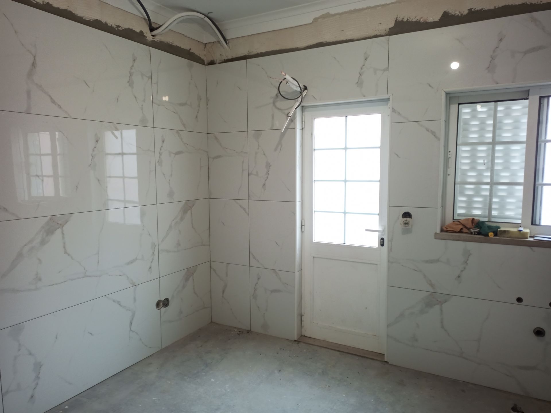 Quarto interior em reforma, com paredes revestidas com azulejos brancos imitando mármore e porta e janela brancas. Fiação elétrica visível.