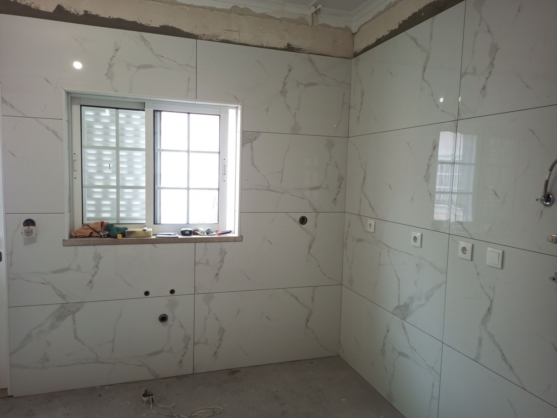 Interior de um quarto com paredes revestidas com azulejos brancos que imitam mármore e uma janela com persianas. Tomadas e luminárias elétricas são visíveis.
