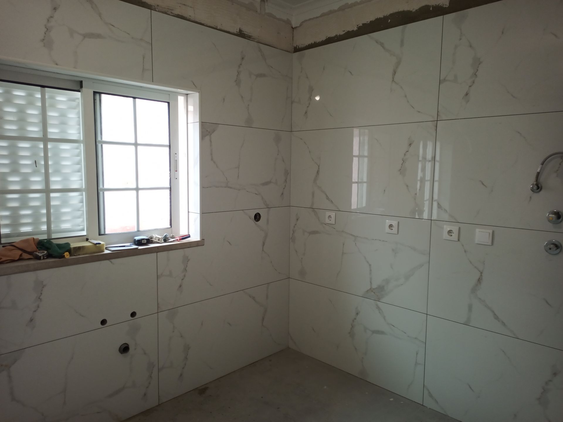 Interior de um quarto com paredes revestidas com azulejos brancos que imitam mármore, uma janela e instalações elétricas.