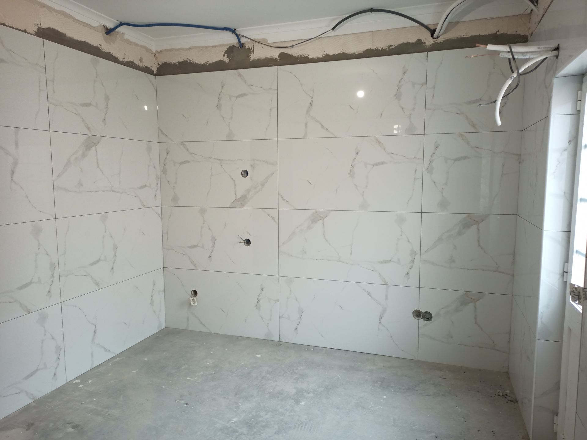 Quarto com azulejos brancos imitando mármore parcialmente instalados nas paredes; piso inacabado e fiação exposta.