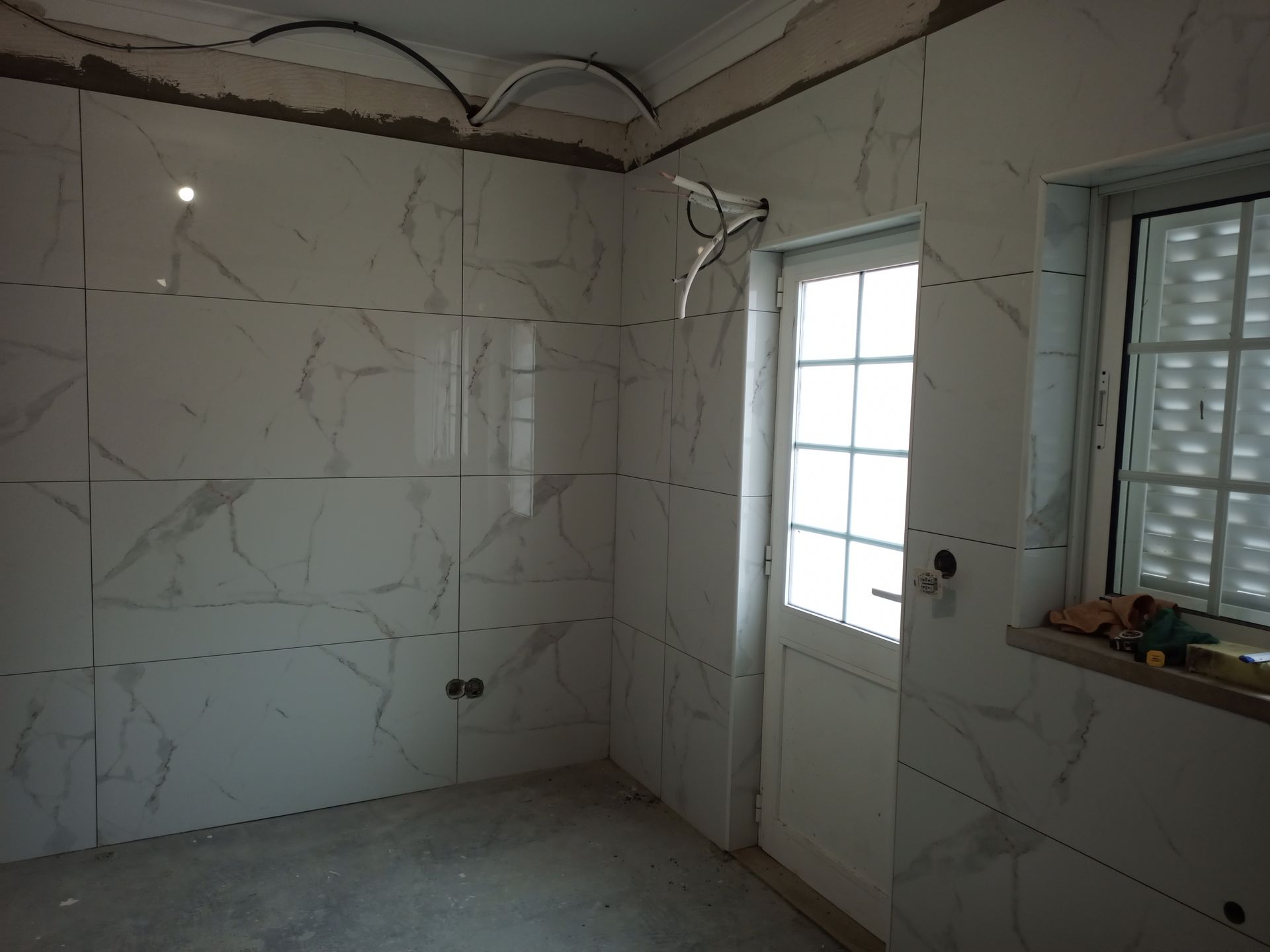 Quarto interior com azulejos brancos imitando mármore nas paredes, porta e janela brancas, fiação elétrica inacabada e piso de concreto.