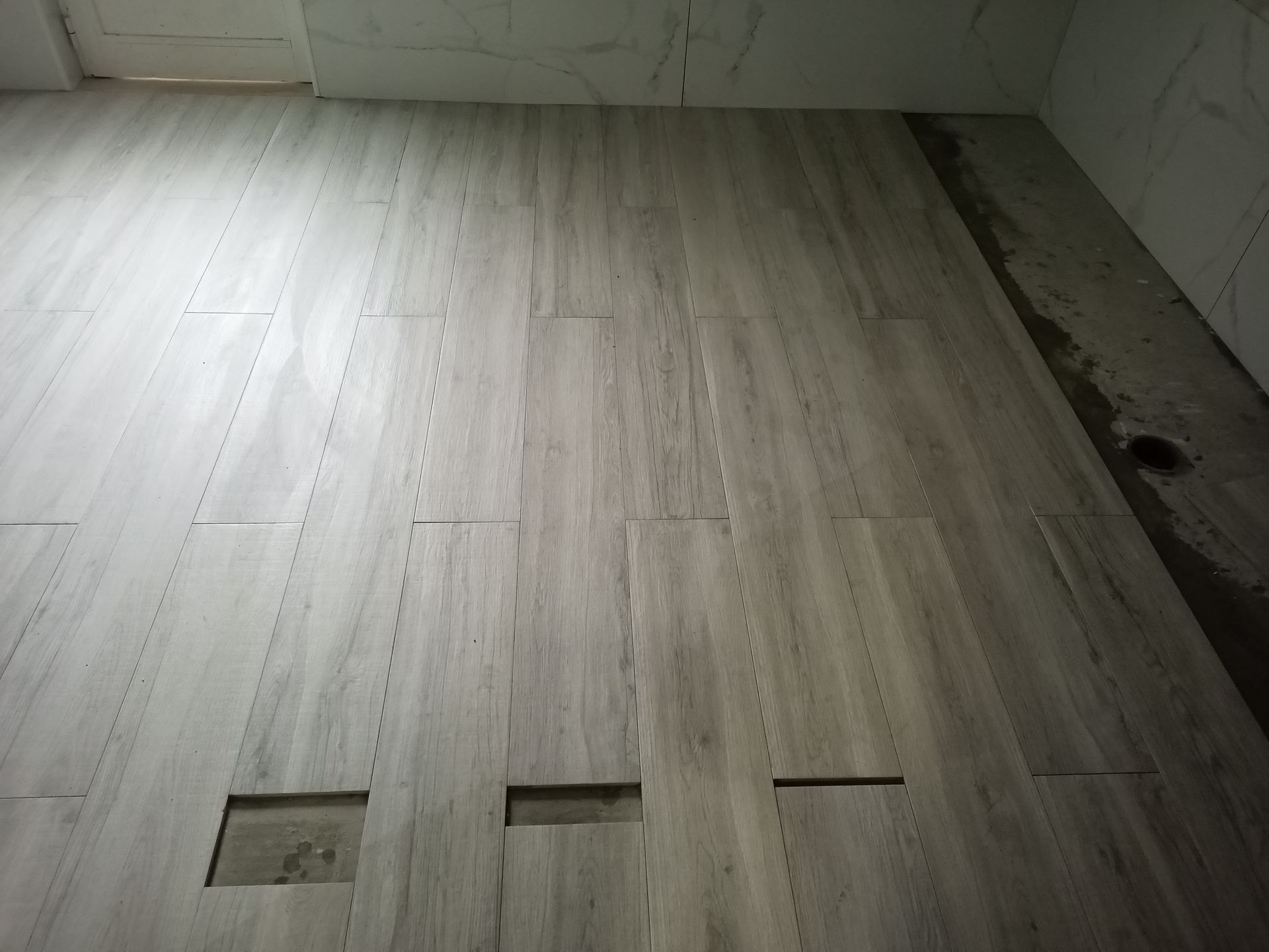 Piso imitando madeira cinza com vários orifícios retangulares, possivelmente para passagem de encanamento ou fiação elétrica, em um cômodo inacabado.