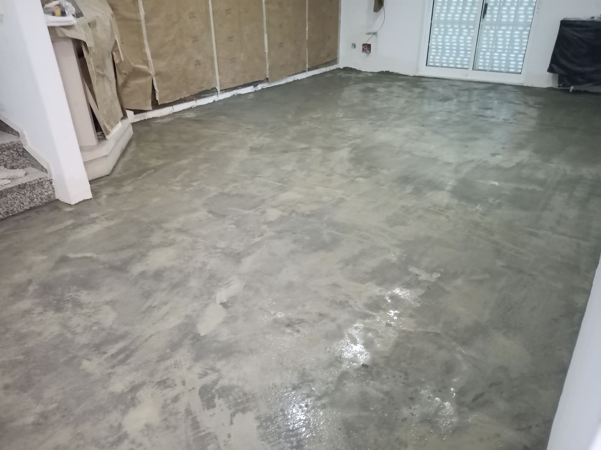 Piso de concreto em um cômodo, parcialmente restaurado com manchas cinza-claro e uma porta à esquerda.