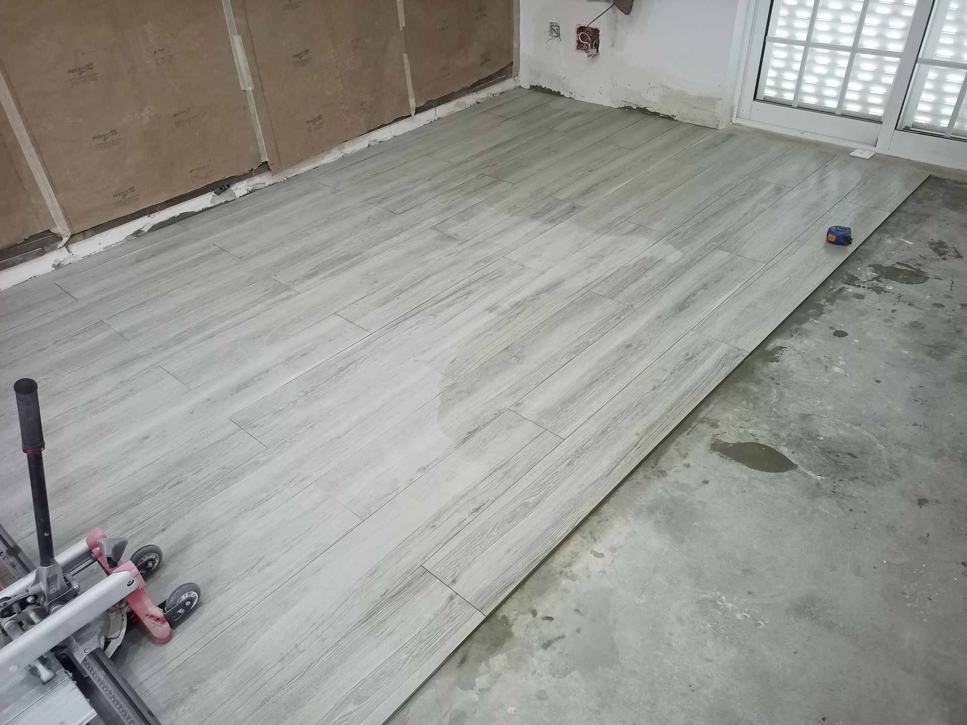 Instalação de azulejos cinza em andamento sobre piso de concreto próximo a uma porta de vidro deslizante.