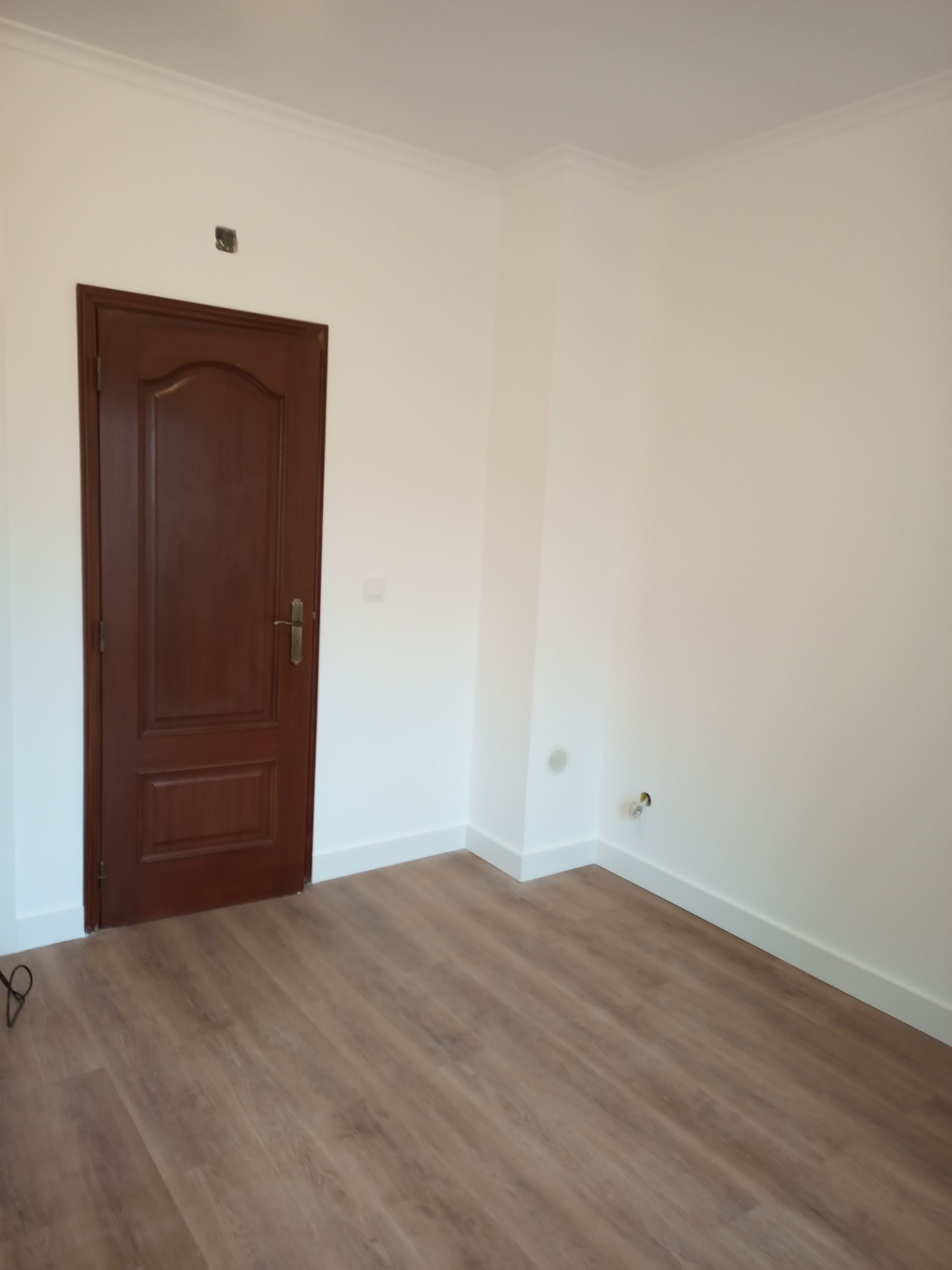Quarto vazio com piso de madeira, paredes brancas, porta marrom e molduras brancas.