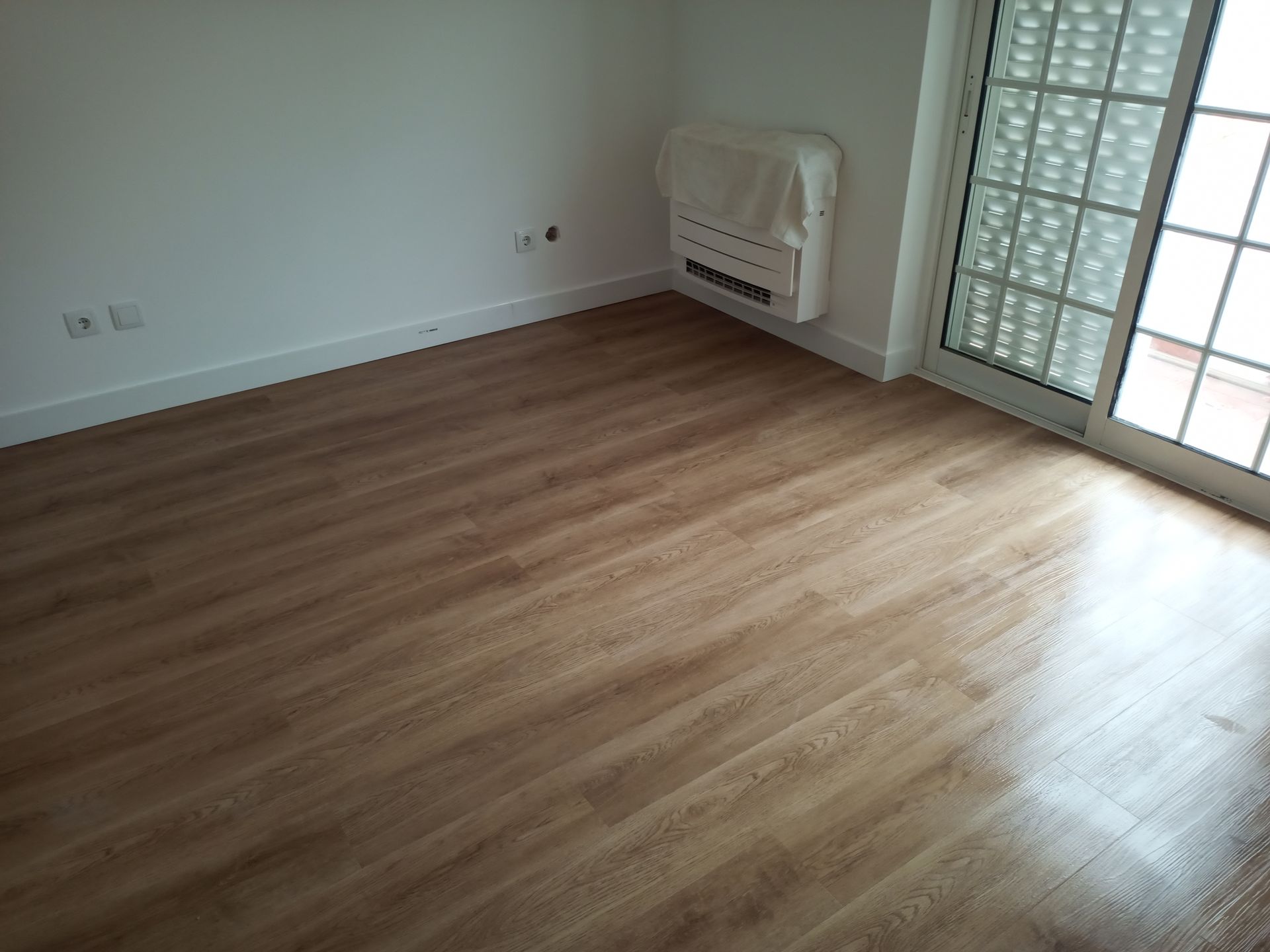 Quarto com piso imitando madeira, paredes brancas, aquecedor e porta de vidro deslizante com grade decorativa.