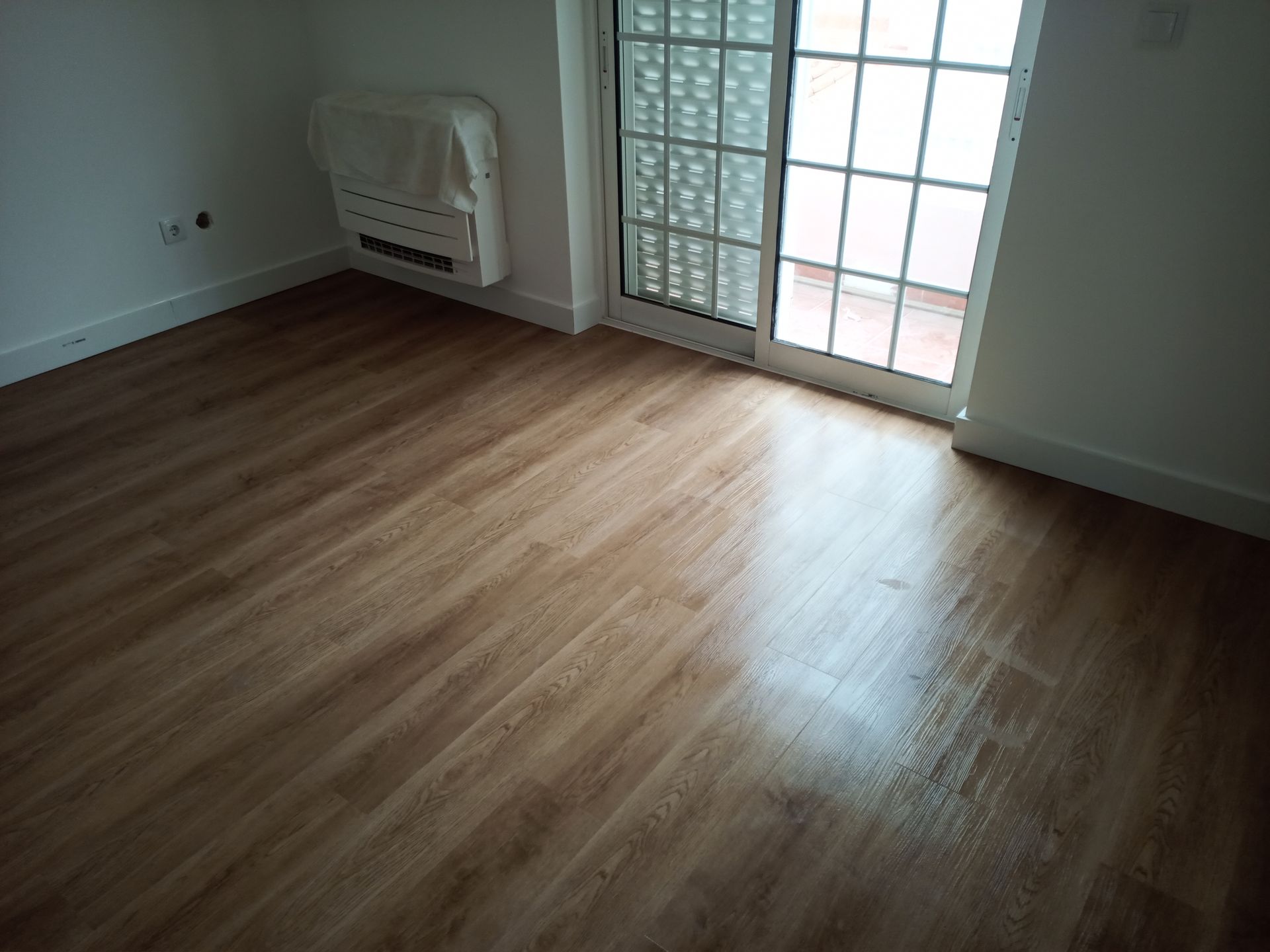 Quarto com piso imitando madeira, paredes brancas, aquecedor e porta de vidro deslizante para uma varanda.